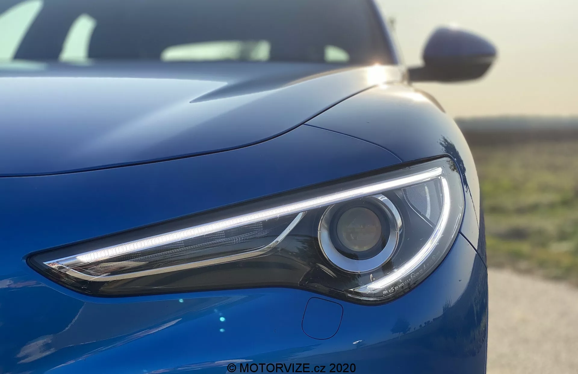 Vista de cerca de la sección frontal izquierda del Alfa Romeo Stelvio 2017 destacando el faro, tomada desde un ángulo bajo. La imagen enfatiza el diseño elegante y la tecnología LED del conjunto de luces del modelo de lujo deportivo con una carrocería de alto brillo en un color azul metálico profundo que refleja un cielo despejado.