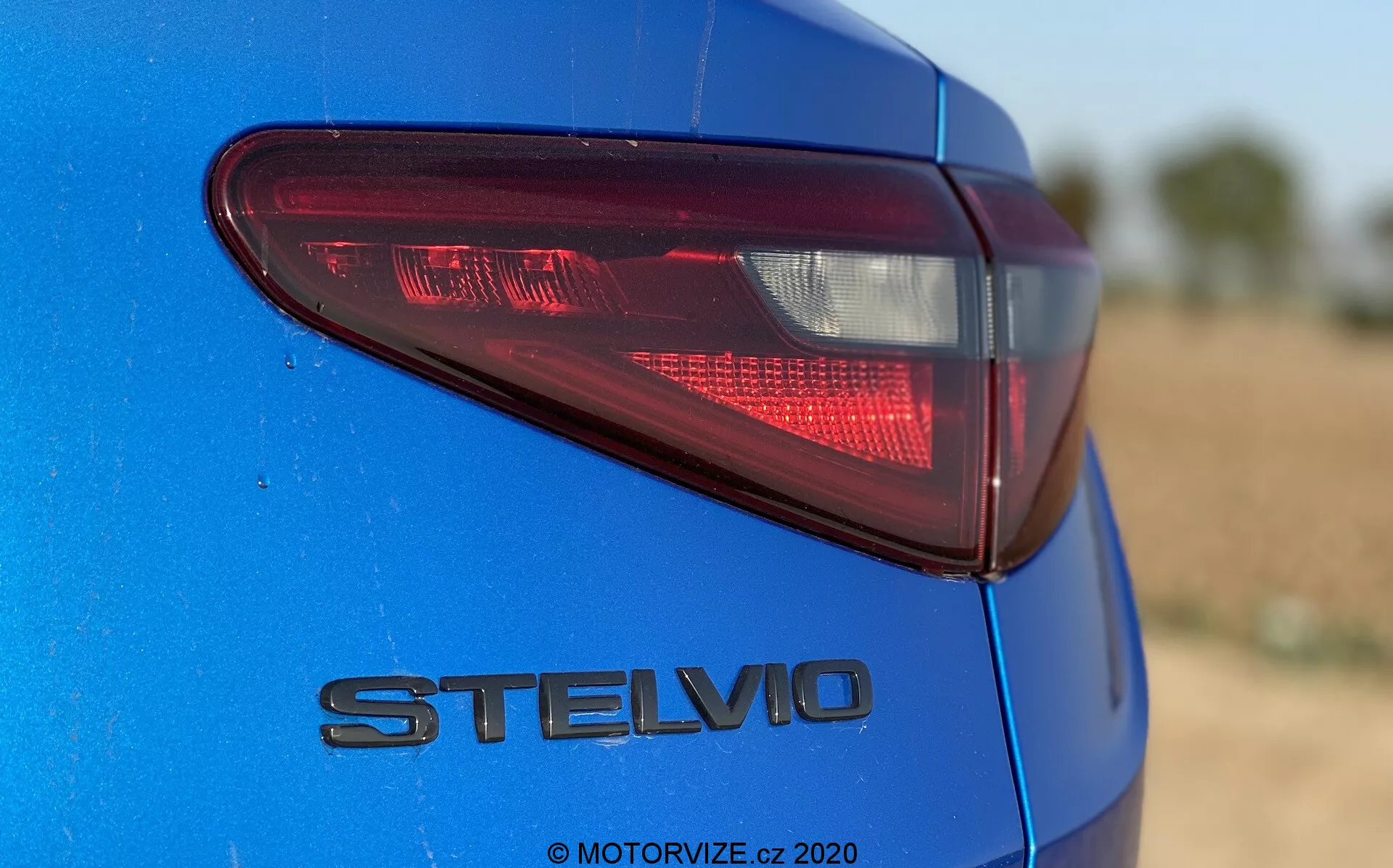 Detalle de la sección trasera del Alfa Romeo Stelvio 2017 azul, capturado desde un ángulo bajo y ligeramente fuera del centro enfocando el lado trasero izquierdo. Destaca la luz trasera LED moderna y el emblema cromado 'STELVIO', mostrando el característico acabado de pintura metálica. La poca profundidad de campo mantiene la luz trasera y el emblema enfocados contra un fondo borroso. La luz natural del sol durante el día mejora la profundidad del color y los reflejos.