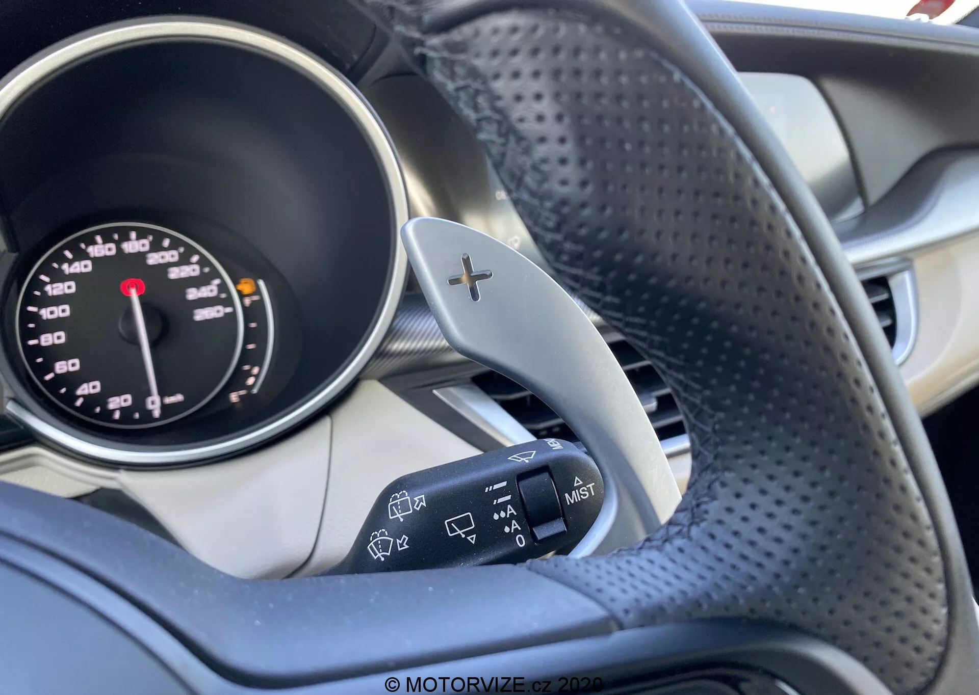 La imagen destaca el interior de un Alfa Romeo Stelvio 2017, enfocándose en el lado del conductor con énfasis en el volante y parte del cuadro de instrumentos. La fotografía está tomada desde el lado derecho del volante, mostrando la porción izquierda del volante, con textura de cuero perforado y un acento metálico con un símbolo de '+' que indica paletas de cambio. También se ve el velocímetro analógico con marcas en mph y km/h y la palanca de señal de giro a la izquierda con controles adicionales para los faros. El fondo presenta elementos interiores borrosos con una mezcla de tonos oscuros y claros. El enfoque detallado en estos controles resalta la funcionalidad de dirección y de iluminación del vehículo.