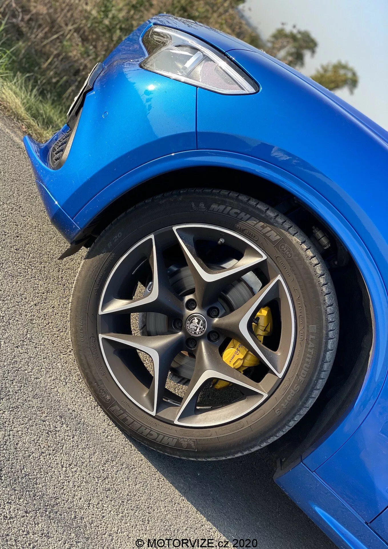 Vista en primer plano de la rueda delantera izquierda y el guardabarros del Alfa Romeo Stelvio 2017 desde un ángulo bajo, enfocándose en la llanta de aleación multirradio de acabado oscuro, el gran disco de freno y la pinza de freno amarilla brillante. Se destacan las líneas y contornos dinámicos de la carrocería, la pintura azul brillante y los neumáticos de alto rendimiento de perfil bajo con letras blancas. Foto diurna que muestra un Alfa Romeo Stelvio deportivo y bien mantenido en condiciones meteorológicas despejadas; marca de agua que dice 'MOTORWIZE.cz 2020'.