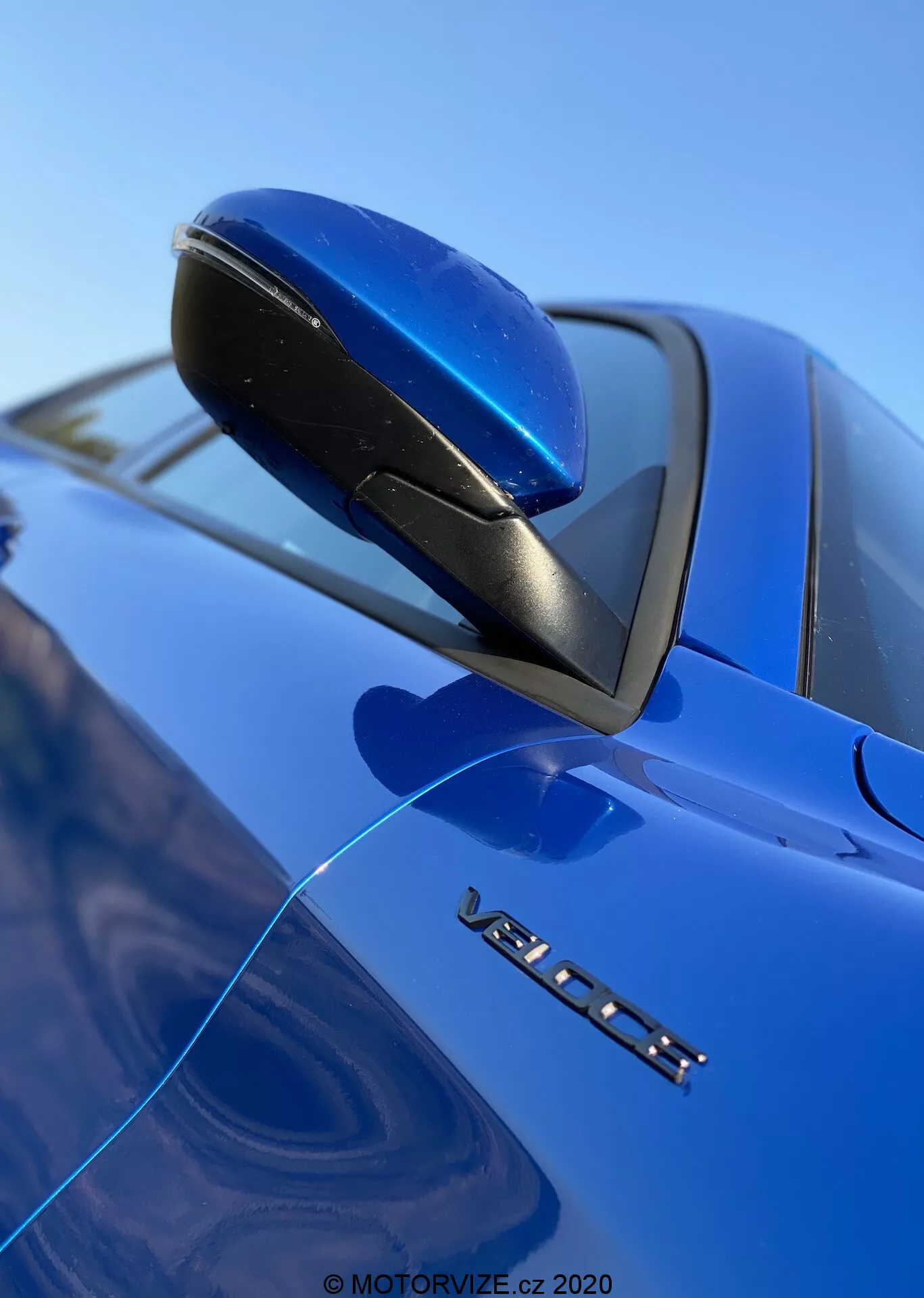 Vista en primer plano y desde un ángulo bajo del espejo lateral de un Alfa Romeo Stelvio 2017 azul, destacando su diseño aerodinámico, base negra e indicador de señal de giro integrado. La marca VOLVO y una insignia de modelo parcial son visibles debajo del espejo, con un enfoque nítido en estos detalles en contraste con un fondo difuminado de parabrisas y cielo, resaltando el acabado de pintura brillante del coche.
