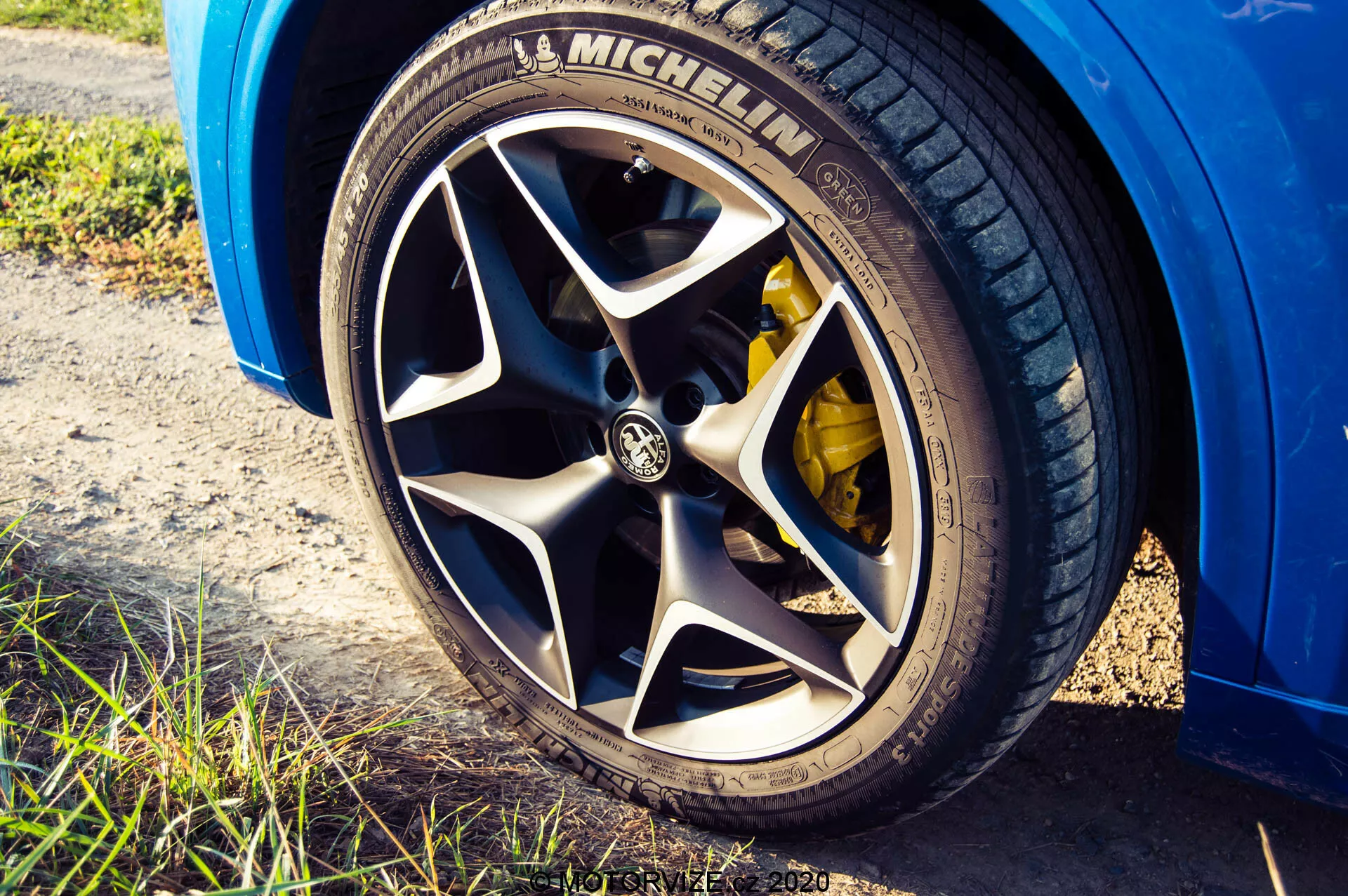 Imagen de perspectiva en ángulo bajo de cerca de la rueda de un Alfa Romeo Stelvio 2017 que muestra un neumático Michelin con un patrón de banda de rodadura visible, llanta de aleación de múltiples radios con el logotipo de la marca, pinza de freno de alto rendimiento amarilla, exterior del coche de color azul brillante, ambientada en luz natural con un toque de entorno exterior y poca profundidad de campo que enfatiza la rueda.