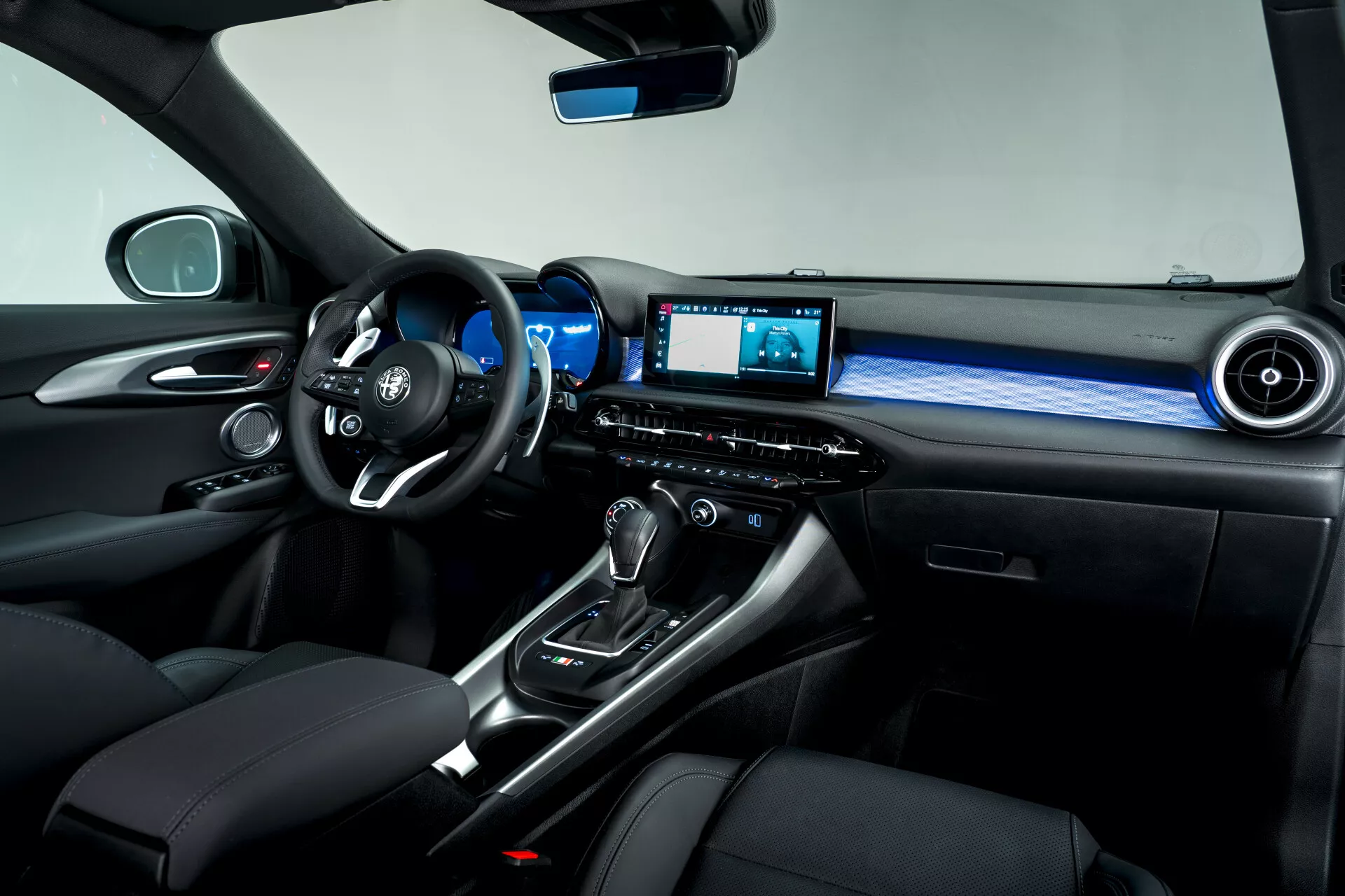 Vista interior del lado del pasajero del Alfa Romeo Tonale 2022, destacando el volante multifunción, el tablero digital, la palanca de cambios de transmisión automática con bota de cuero, selector de modo de conducción, sistema de infotenimiento con pantalla, iluminación ambiental y asientos de cuero premium con costuras en contraste.