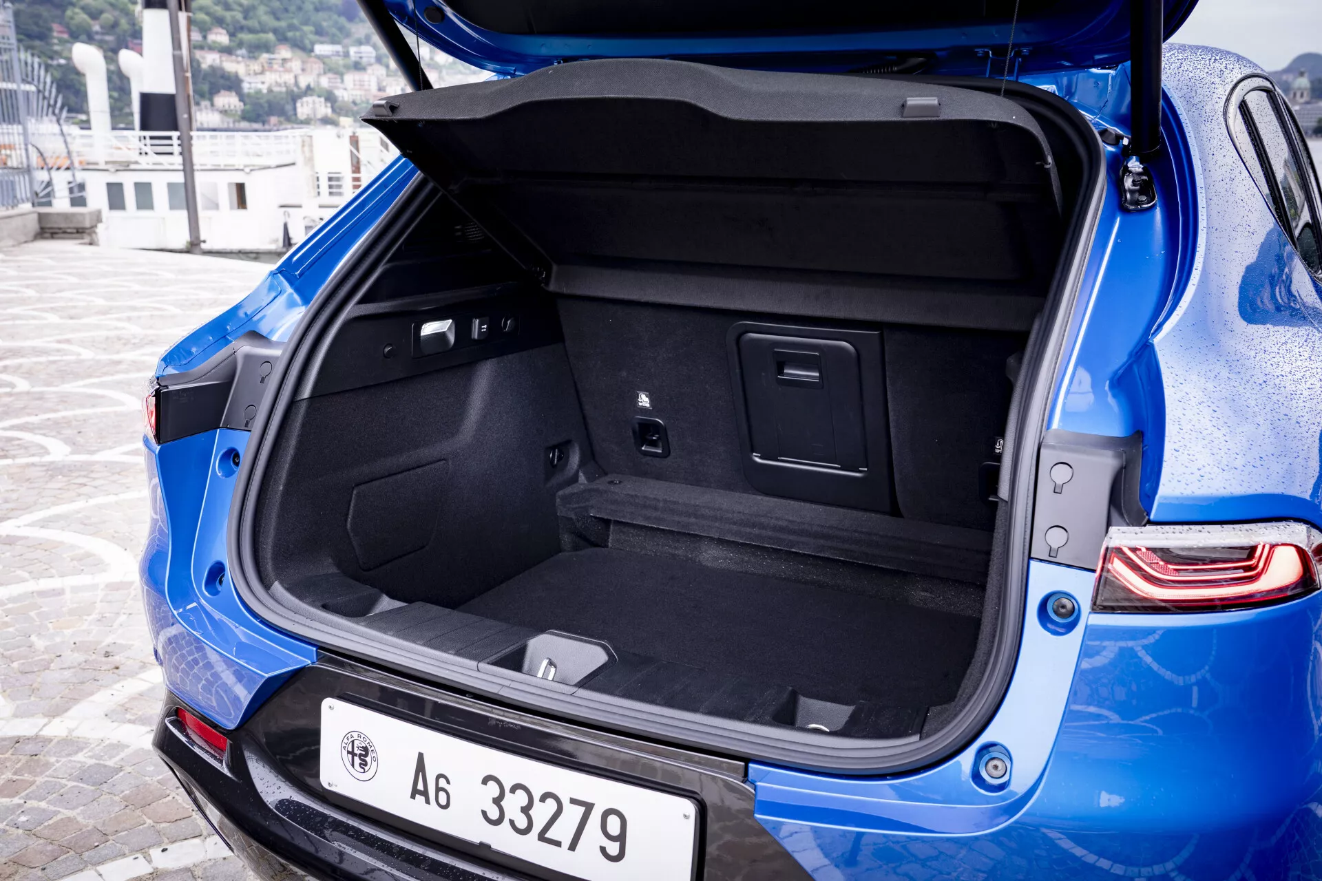Vista angular detallada de la parte trasera del Alfa Romeo Tonale 2022 azul presentando el espacio abierto del maletero con interior alfombrado, compartimentos integrados y una toma de corriente, junto con el parachoques trasero visible con sensores de estacionamiento y luces traseras LED modernas.