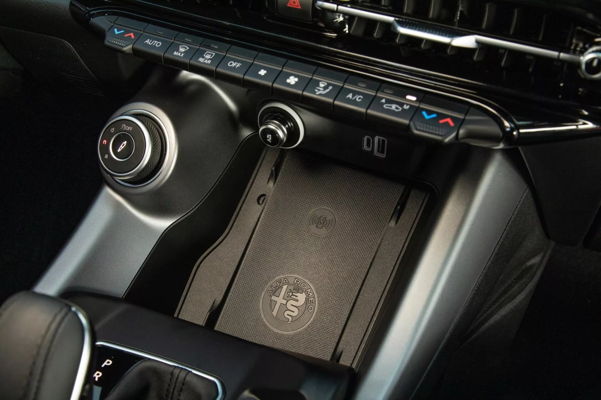 Vista detallada del centro de control de Alfa Romeo Tonale 2022, mostrando panel de control climático, almohadilla de carga inalámbrica, palanca de cambios y dial de control de infoentretenimiento con acabado premium y diseño moderno.