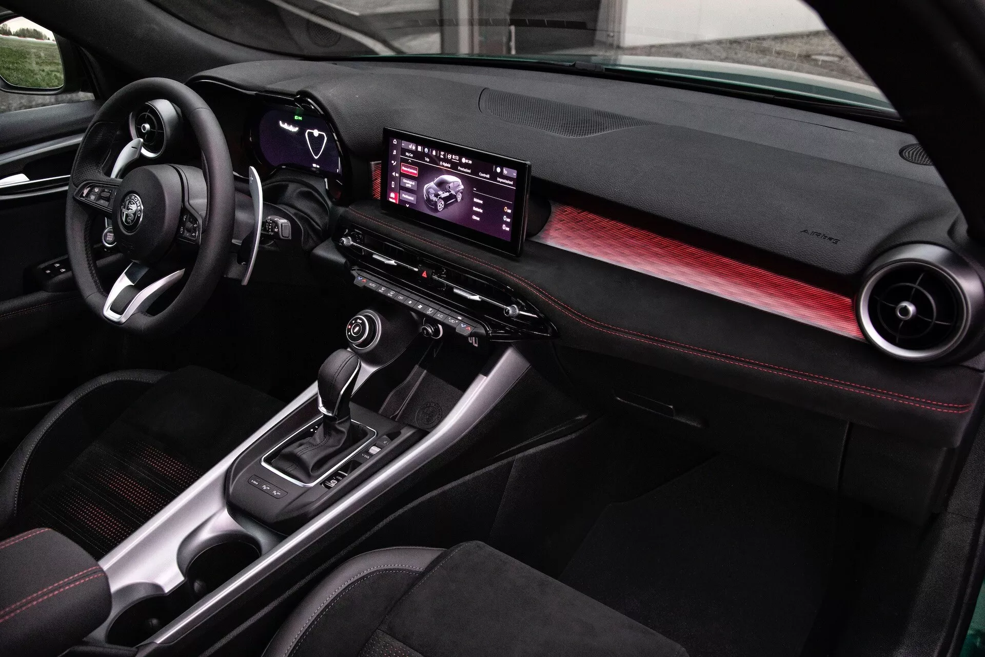 Interior del Alfa Romeo Tonale 2022 con un tablero moderno, centro de infotainment digital, detalles en fibra de carbono y asientos con costuras rojas, visto desde la perspectiva del pasajero delantero