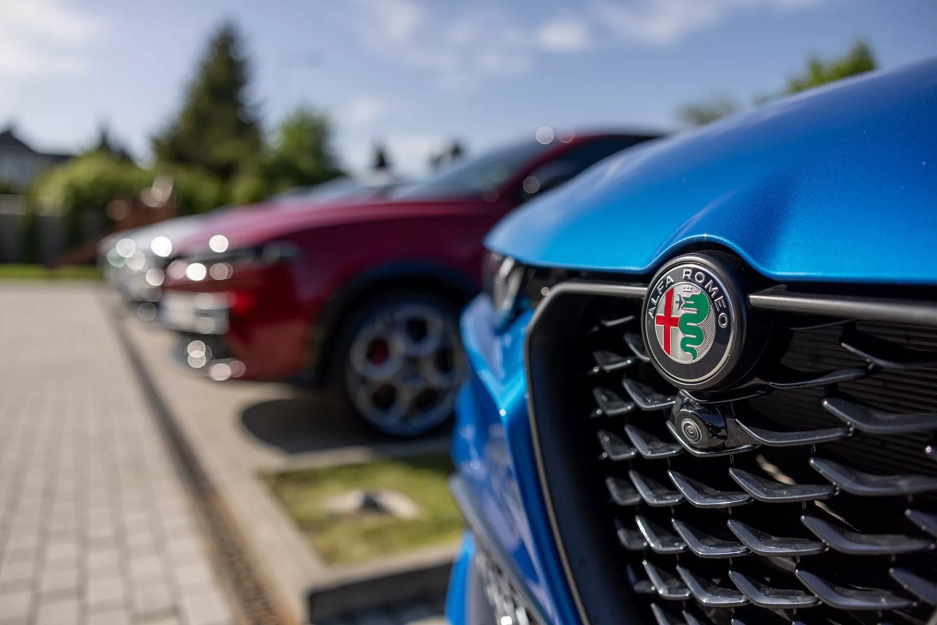 Vista de cerca de la parrilla frontal del Alfa Romeo Tonale 2022 azul con enfoque en el emblema, con lamas verticales y un fondo desenfocado de otros vehículos