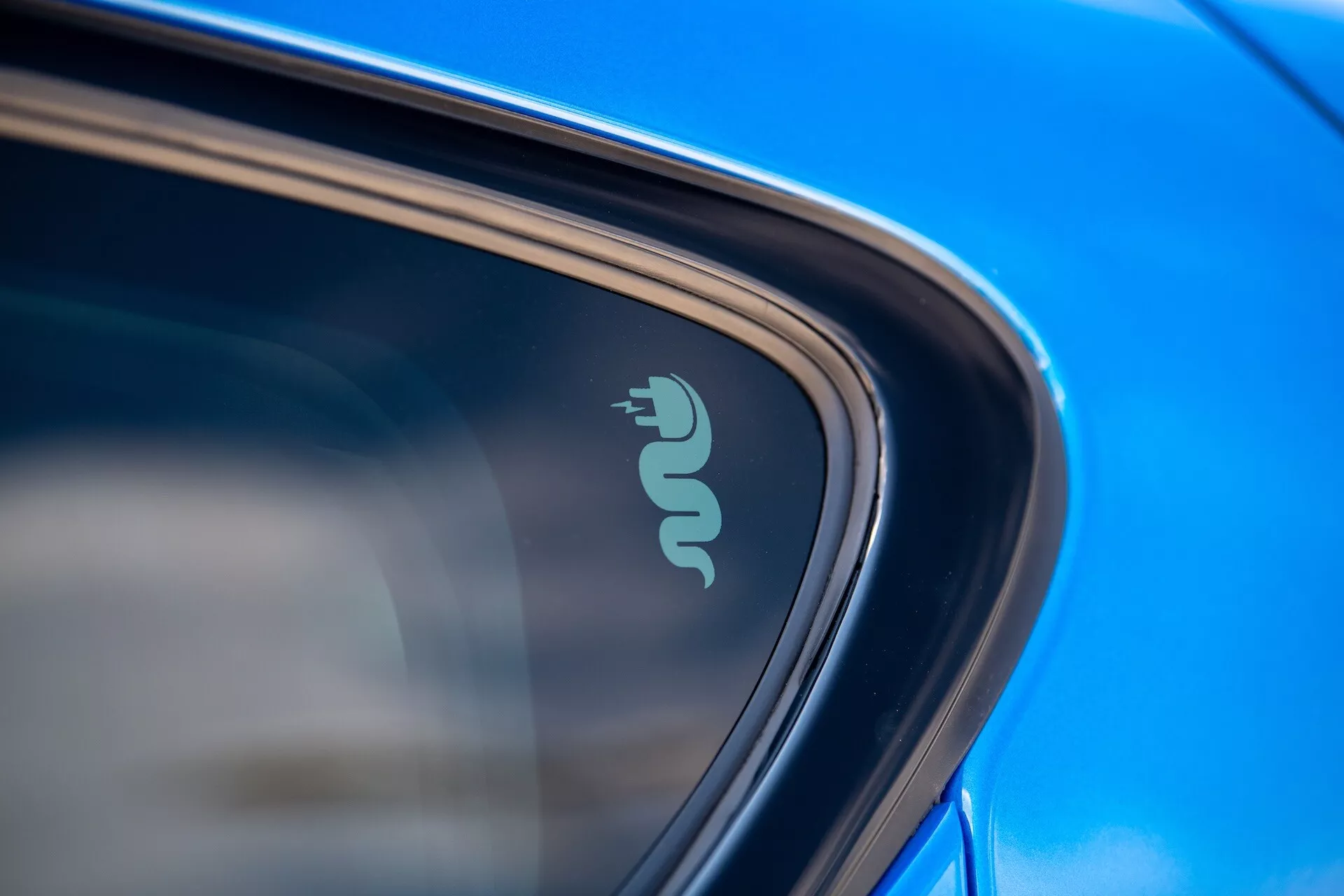 Detalle de un primer plano del Alfa Romeo Tonale 2022 mostrando una insignia tecnológica que indica capacidad eléctrica o híbrida cerca de la ventana del coche, resaltada por pintura azul reflectante y un diseño elegante a la luz natural del día