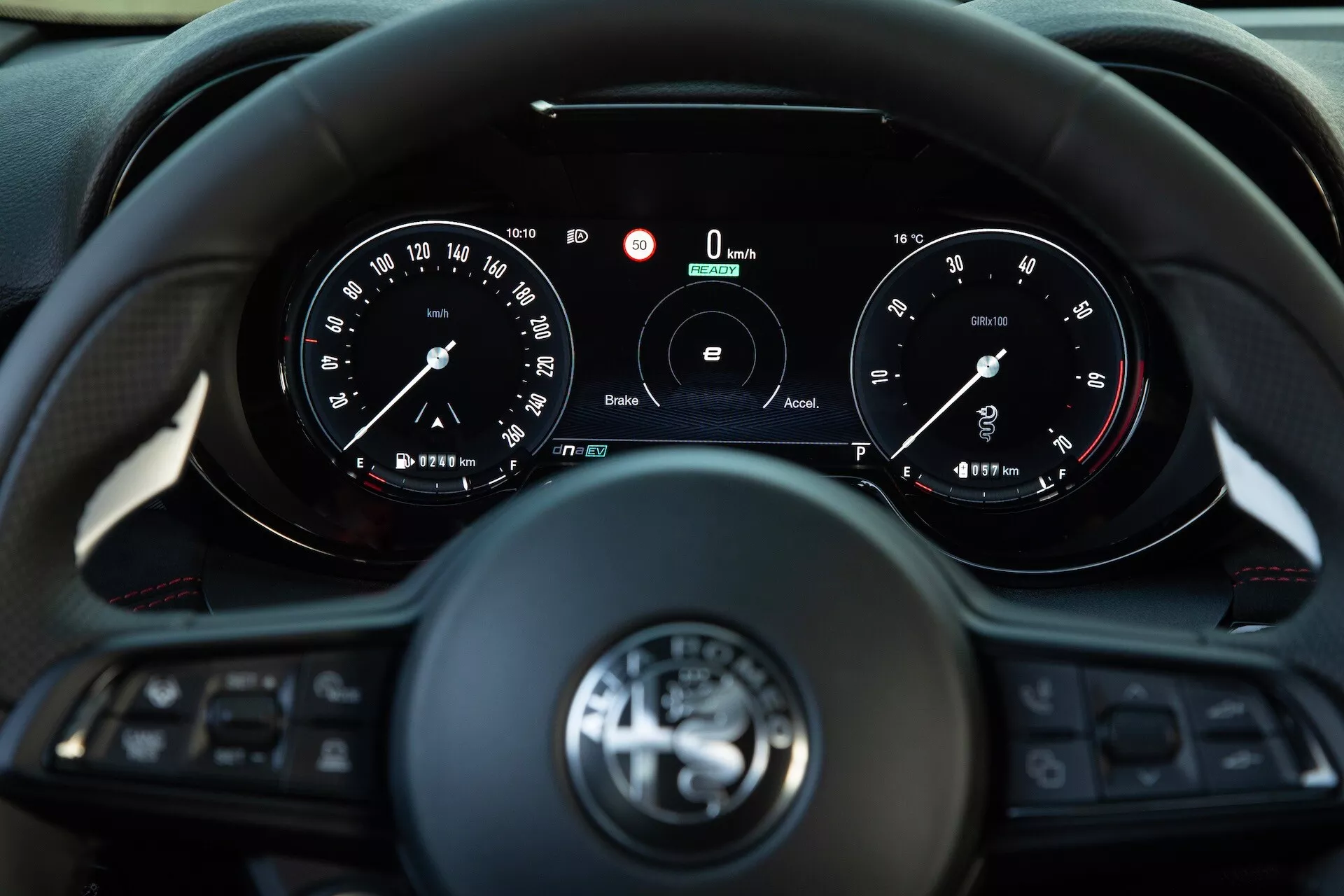 Interior del Alfa Romeo Tonale 2022 mostrando la vista del conductor de un volante multifuncional de tres radios e instrumentos con medidores análogos y una pantalla digital central que indica velocidad y RPM cero, con tecnologías de conectividad y asistencia al conductor