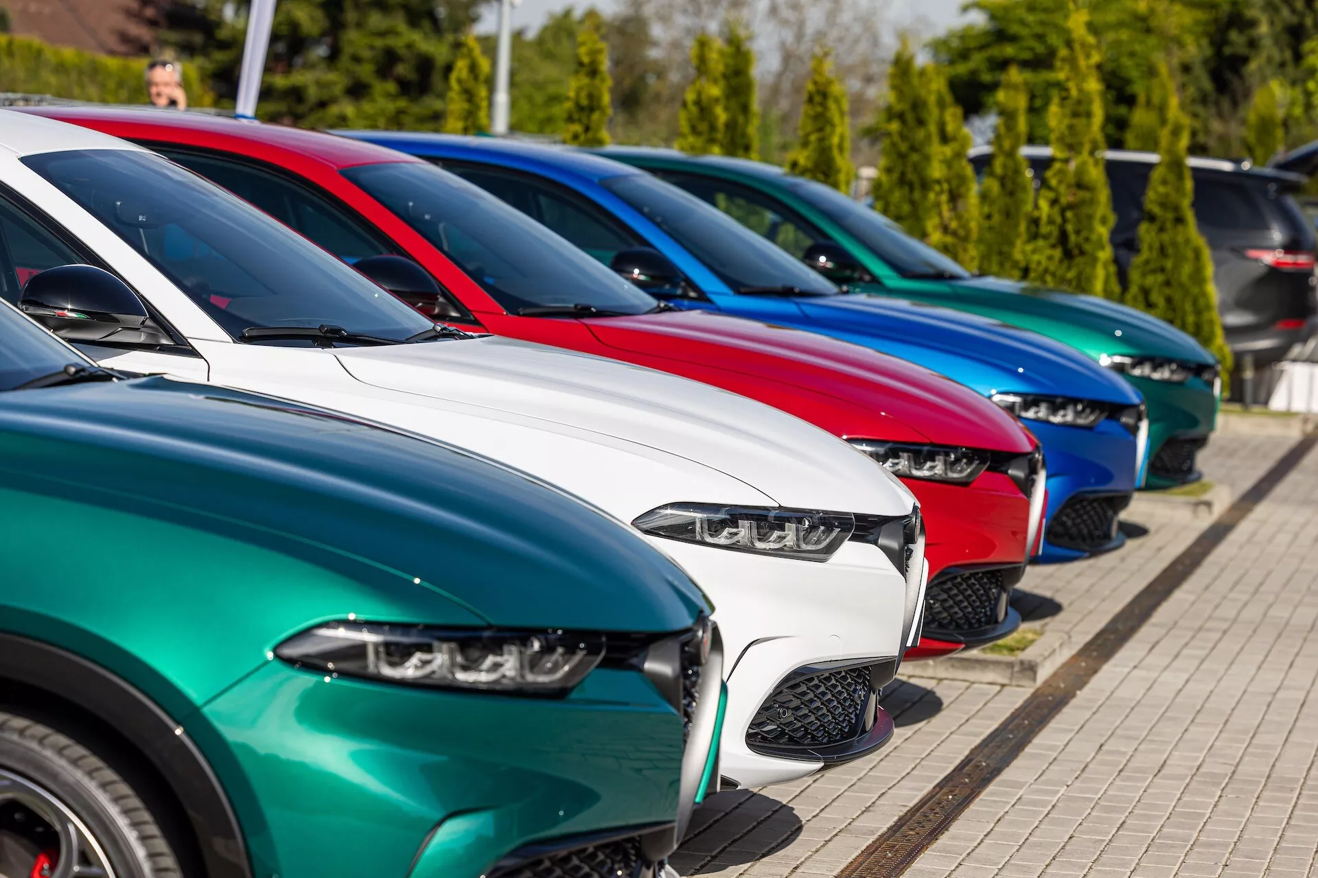 Fila de modelos Alfa Romeo Tonale 2022 aparcados en diagonal mostrando vista frontal tres cuartos destacando faros LED, diseños de parrilla variados, acabados de pintura brillante y ruedas de aleación bajo luz solar brillante con fondo de vegetación