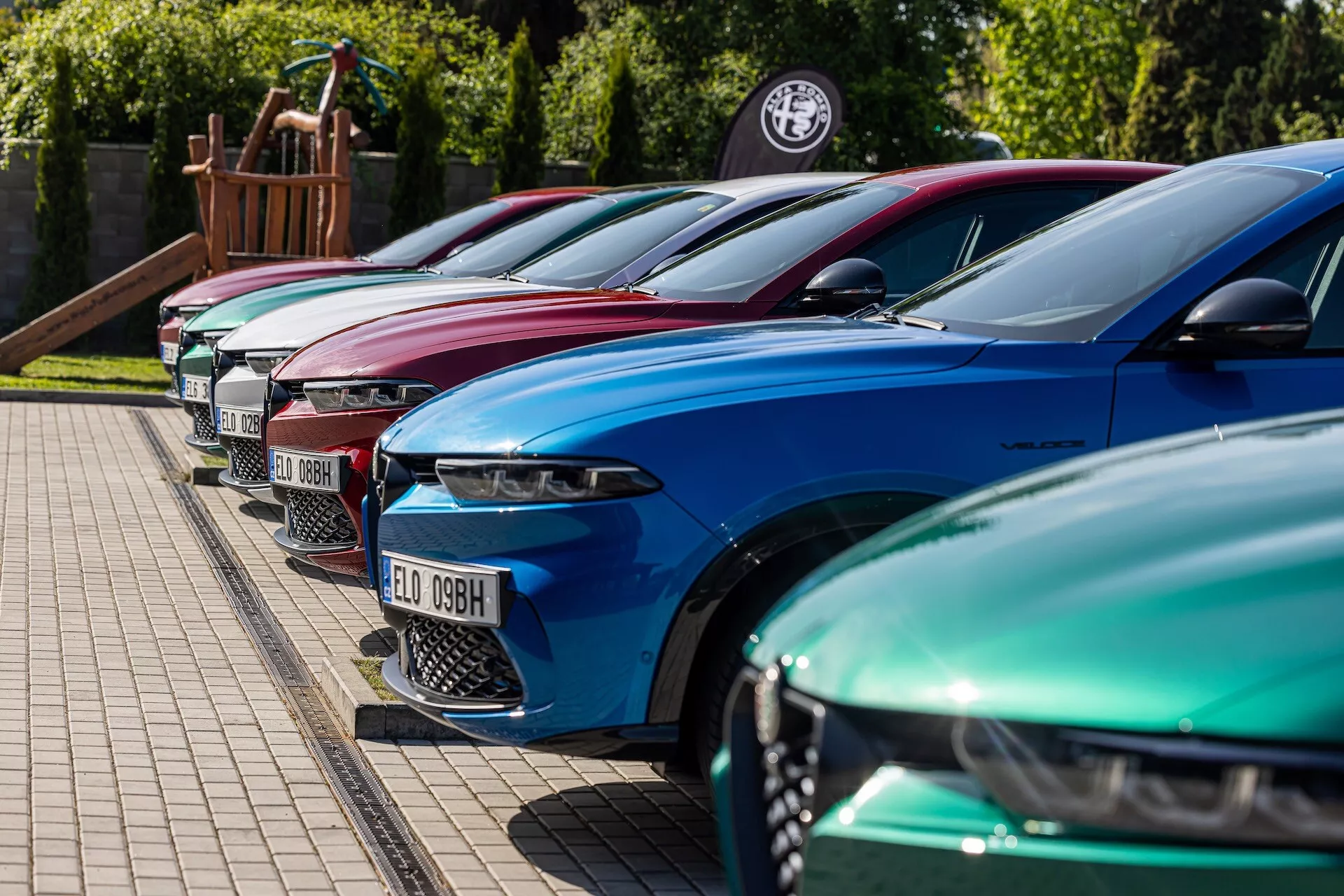 Línea de Alfa-Romeo Tonale 2022 en colores vibrantes mostrando diseño de lujo con faros LED avanzados, estacionados en formación diagonal enfocando el lado frontal izquierdo desde un ángulo de altura baja a media en una área pavimentada al lado de un borde con césped, posiblemente en una concesionaria o evento de entusiastas de autos