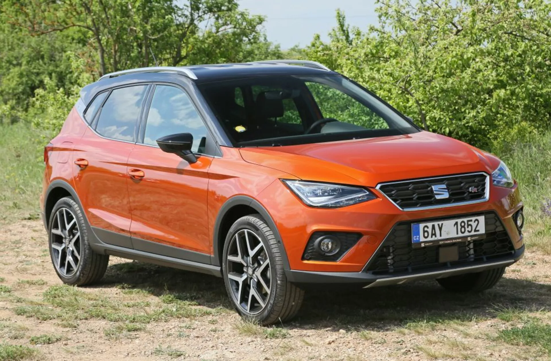 Crossover/SUV naranja aparcado sobre hierba en vista frontal de tres cuartos mostrando luces diurnas LED, diseño moderno y afilado con ruedas de aleación y barras de techo bajo la luz del sol