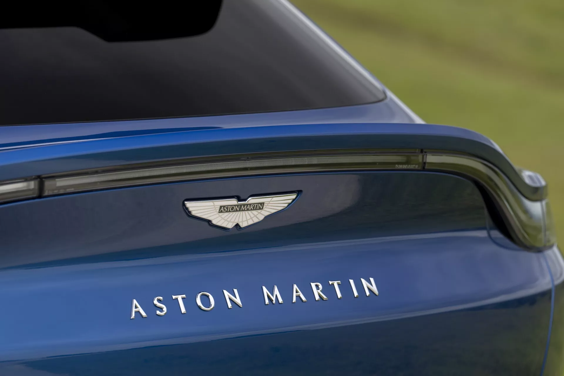 Imagen detallada de la tapa del maletero del Aston Martin DBX 2020, mostrando el nombre y logo de Aston Martin en plata, con enfoque en el diseño elegante, deportivo y el lujoso exterior azul metálico del coche