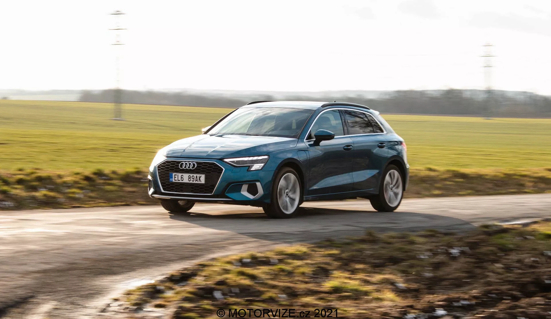 Toma dinámica de un hatchback moderno, probablemente el Audi A3 Sportback 2020, capturado en movimiento. La imagen está tomada desde un ángulo ligeramente bajo, enfatizando la altura del coche y creando una sensación dinámica. Muestra una vista frontal de tres cuartos con el vehículo moviéndose de derecha a izquierda. Entre las características de diseño notables se incluyen faros LED elegantes, una gran parrilla frontal, ruedas de aleación grandes, espejos laterales aerodinámicos con posibles señales integradas y un acabado de pintura metálica con líneas de contorno. El fondo está desenfocado, lo que mejora la sensación de velocidad, mientras que el coche en sí mantiene un enfoque nítido, destacando su diseño contemporáneo y estética premium.