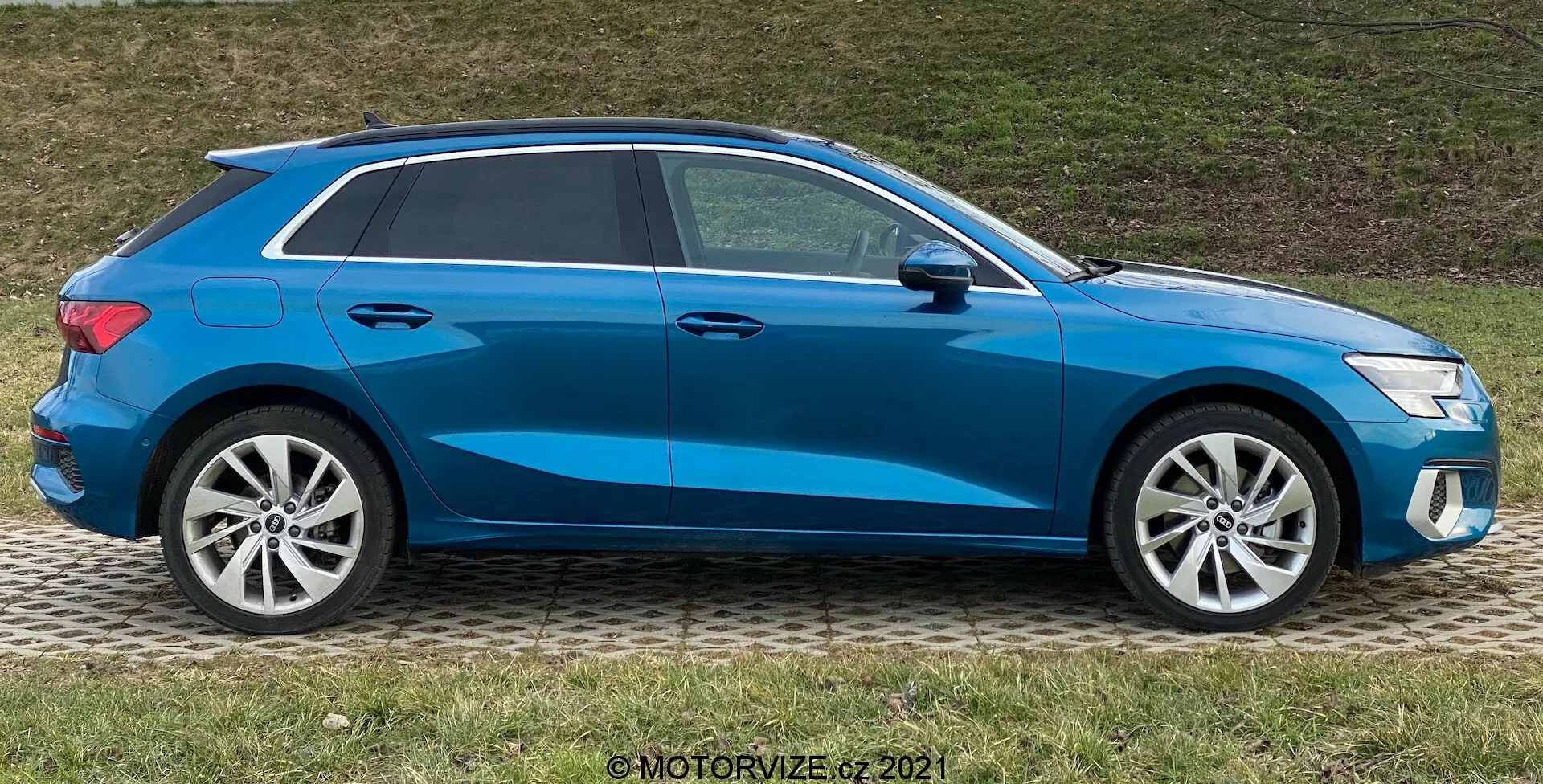 Vista lateral de un Audi A3 Sportback azul 2020, que muestra su diseño moderno de hatchback con una línea de techo inclinada deportiva, llantas de aleación, ventanas cerradas y espejos laterales del color de la carrocería con señales de giro integradas, estacionado sobre el césped junto a una acera.