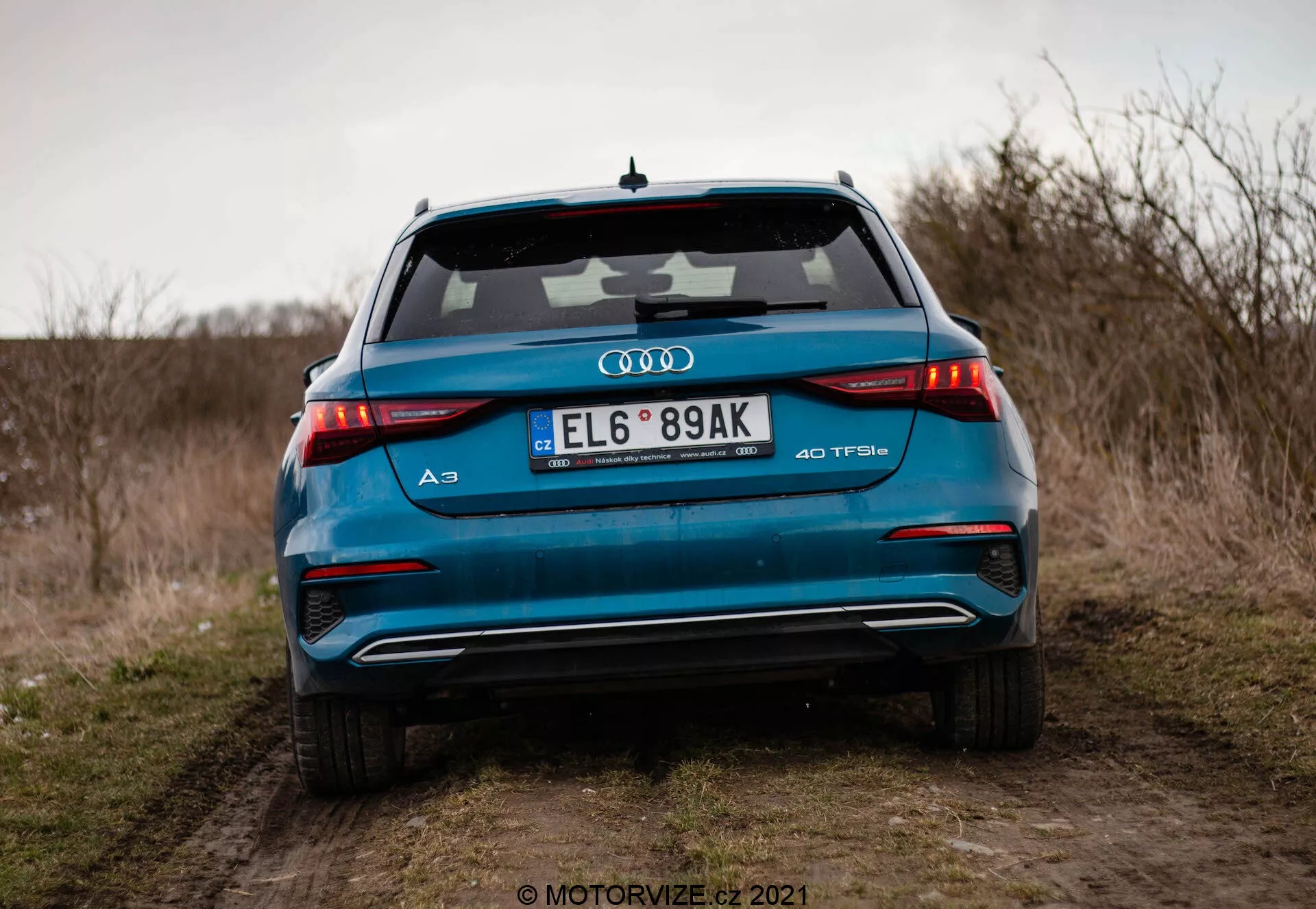 Vista detallada de la parte trasera de un vibrante Audi A3 Sportback 40 TFSI azul de 2020, destacando sus avanzadas características de diseño como las luces traseras LED horizontales, los tubos de escape dobles, el emblema del motor turboalimentado TFSI, el alerón integrado y la antena de aleta de tiburón, fotografiado desde un ángulo bajo en un camino de tierra con un campo abierto y cielo nublado al fondo.