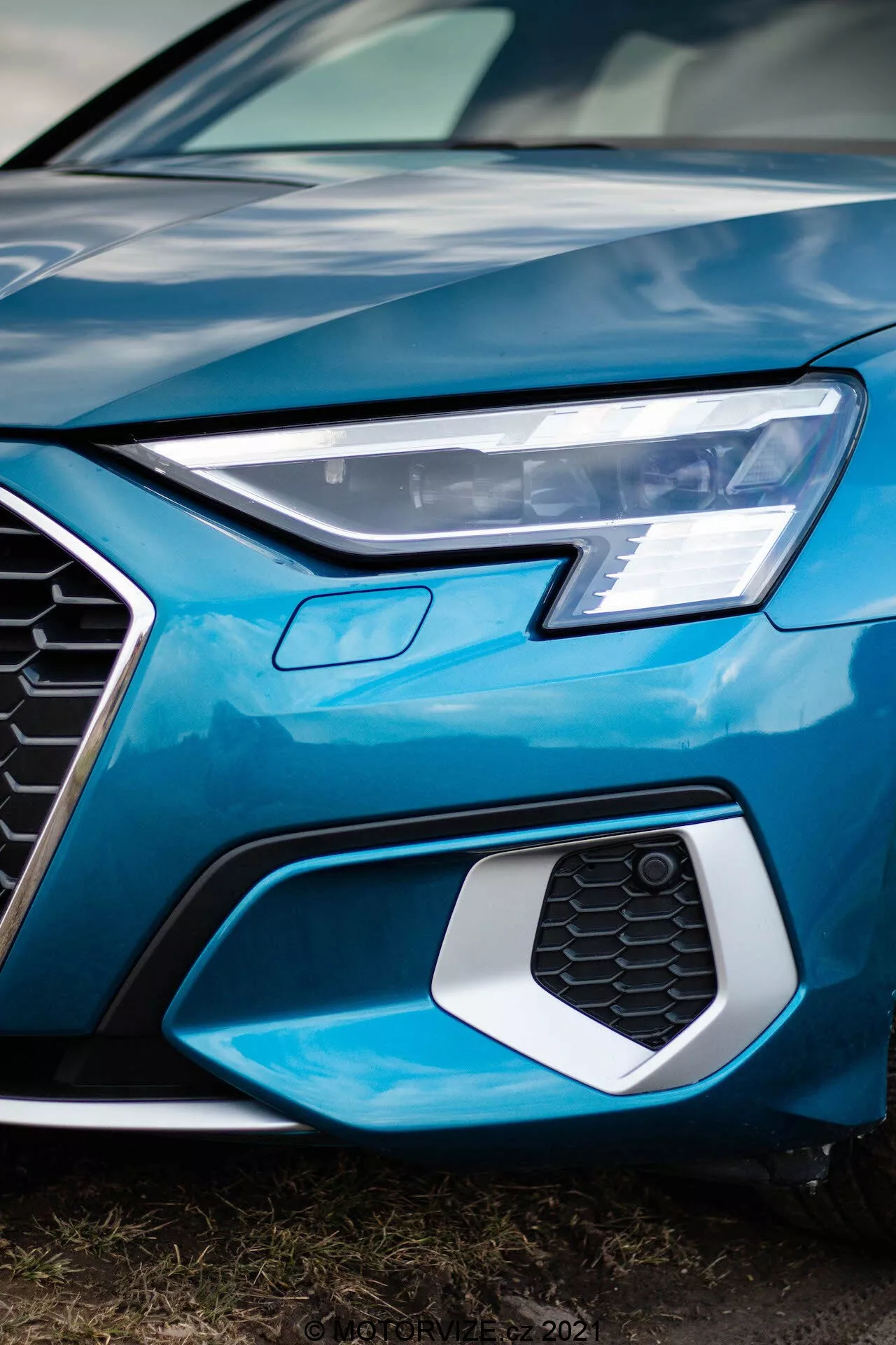 Toma de primer plano en ángulo bajo de la parte delantera izquierda de un Audi A3 Sportback 2020, capturando el faro LED afilado, la parrilla hexagonal con patrón de panal que posiblemente aloja sensores o insignias, y parte del capó y parachoques azul brillante y brillante, enfatizando el diseño moderno y sofisticado y la tecnología automotriz avanzada.