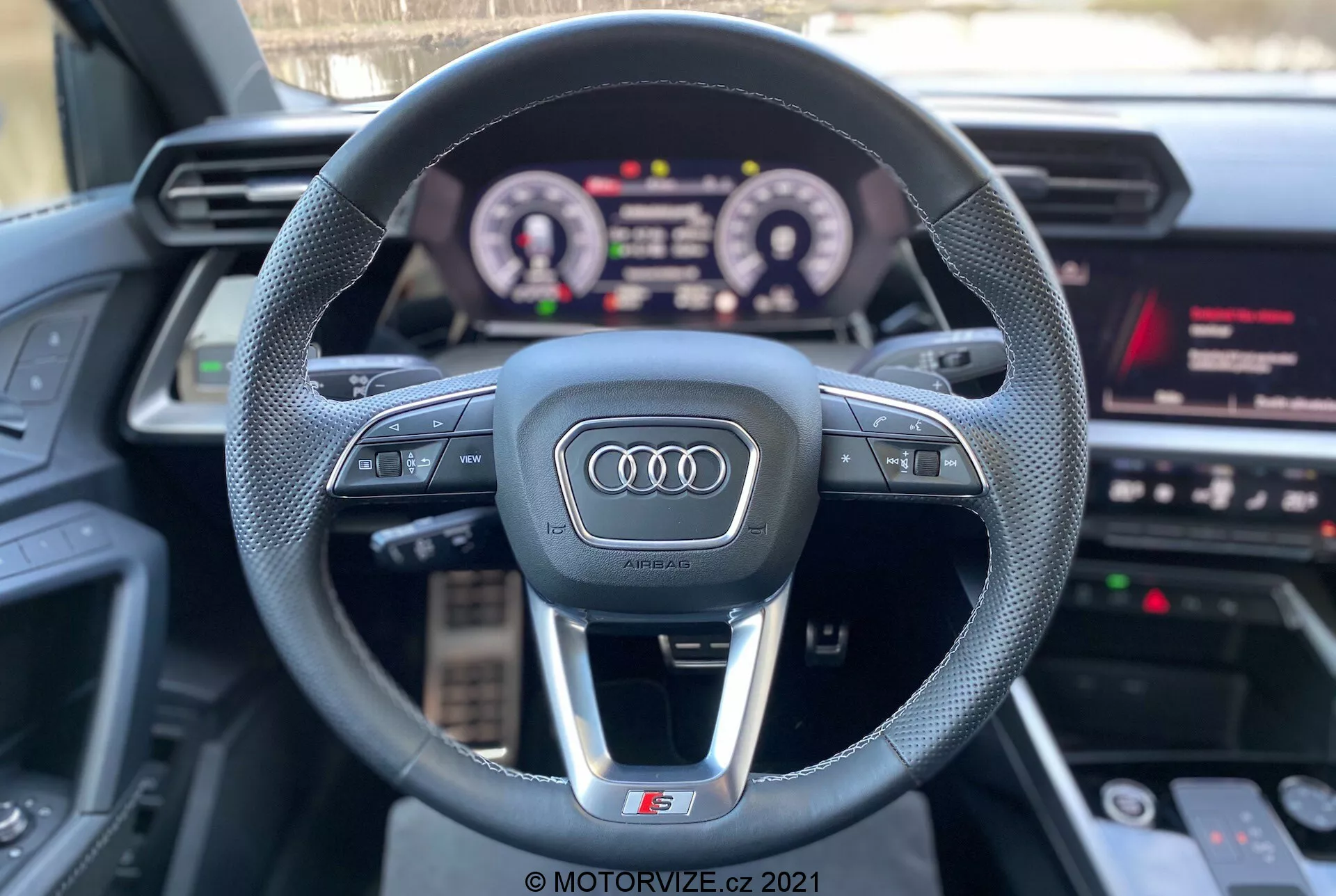 Vista en primera persona desde el asiento del conductor de un Audi A3 2020 (A3 Sportback) que muestra el volante deportivo de tres radios con el logotipo de Audi, botones de control multifunción, tablero de instrumentos digital iluminado y parte del sistema de infoentretenimiento con medios, navegación y configuración del vehículo. Las características resaltan la tecnología interactiva y los componentes de lujo con un enfoque en la experiencia de conducción.