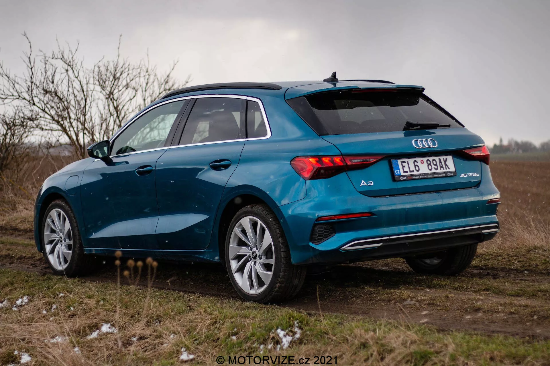 Un Audi A3 2020 (Sportback) azul fotografiado desde un ángulo de tres cuartos trasero en un campo abierto bajo un cielo nublado. La imagen destaca el lado derecho del coche, las luces traseras LED, el sistema de escape doble, las llantas de aleación de varios radios y el alerón trasero. El vehículo está estacionado en una superficie no pavimentada con trozos de hierba y un toque de nieve o escarcha, lo que sugiere condiciones climáticas frías. El ambiente general es sombrío, con un enfoque en el diseño deportivo y premium del Audi A3.