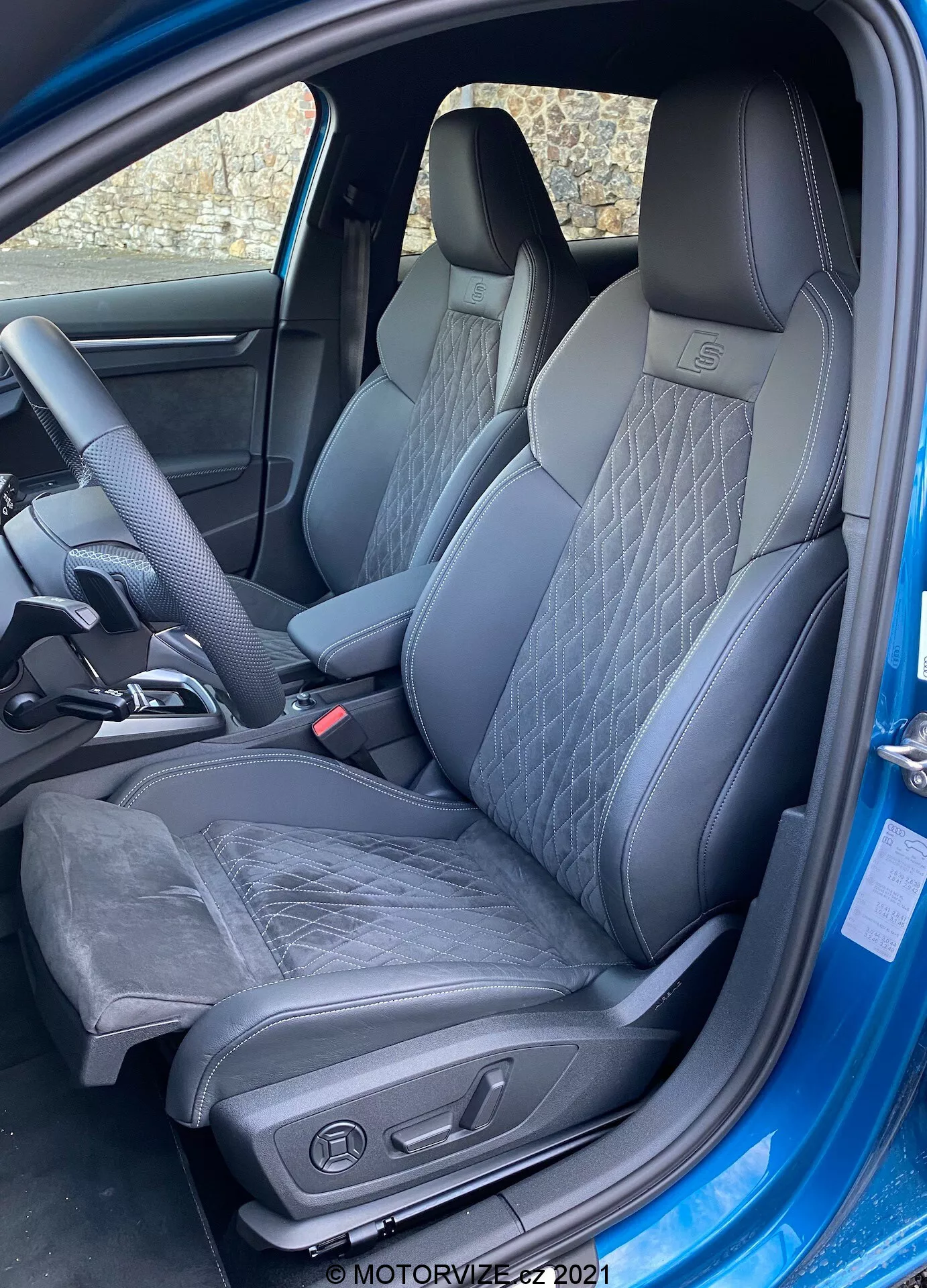 Toma exterior en ángulo medio del interior del Audi A3 Sportback 2020, enfocándose en el asiento del conductor. La puerta está completamente abierta, revelando un asiento deportivo y moderno con cuero y posiblemente Alcantara. El asiento presenta patrones de costura acolchada y refuerzos laterales pronunciados para soporte lateral. Son visibles los controles de ajuste eléctrico del asiento, que sugieren ajustes de altura, inclinación y soporte lumbar. El interior es monocromático con tonos oscuros, contribuyendo a una sensación premium. También es visible una parte del volante ergonómico con área de descanso para el pulgar, lo que indica un volante deportivo.