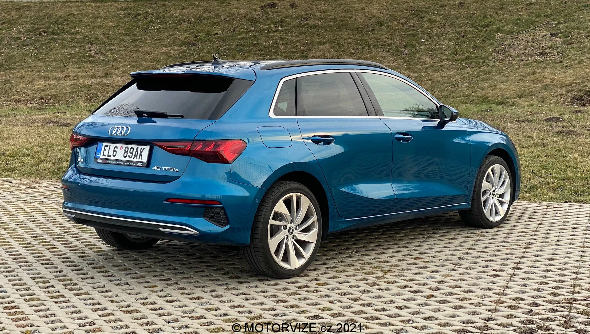 La imagen muestra un Audi A3 2020 Sportback azul desde una vista trasera de tres cuartos, enfatizando el lado trasero derecho. Los detalles clave incluyen llantas de aleación de múltiples radios con acabado plateado, luces traseras LED parcialmente visibles, carrocería aerodinámica con una línea de hombro pronunciada, insignias traseras que indican el modelo y posiblemente el nivel de equipamiento, cristales de privacidad traseros más oscuros, pequeño alerón en el techo, rieles en el techo para versatilidad y salidas de escape dobles en el parachoques trasero inferior. El coche está aparcado sobre una superficie con patrones y un fondo de césped, sugiriendo un entorno no muy urbano.