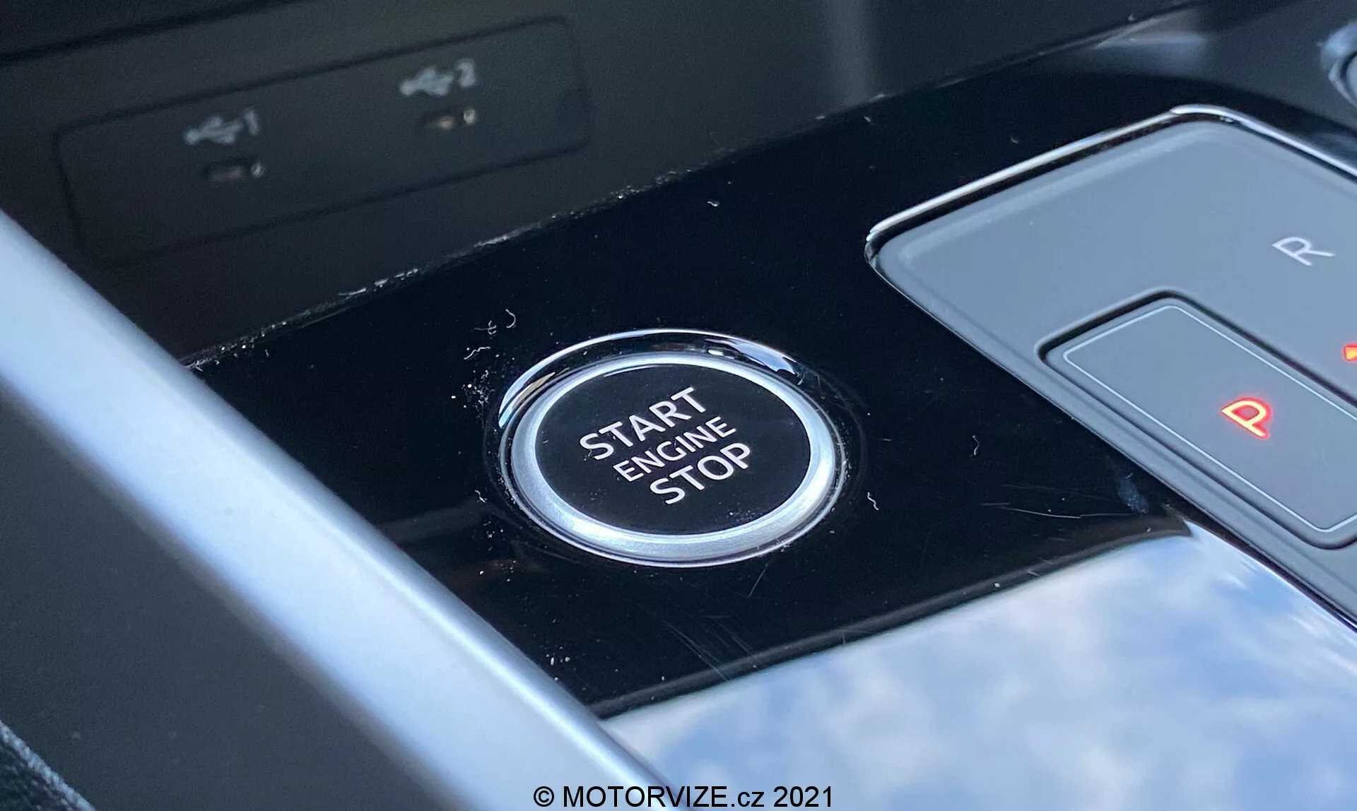 Vista en primer plano del botón de arranque/parada del motor del Audi A3 Sportback 2020 situado en el área de la consola central. El botón muestra prominentemente el texto 'START ENGINE STOP' en letras mayúsculas, indicando su función de encender o apagar el motor. Visto desde la perspectiva del conductor, el panel negro brillante y la palanca de cambios parcialmente visible etiquetada con 'R' y 'P' sugieren una transmisión automática. La poca profundidad de campo enfatiza la claridad del botón contra un fondo ligeramente borroso, apuntando a un interior de vehículo moderno con características tecnológicas avanzadas.