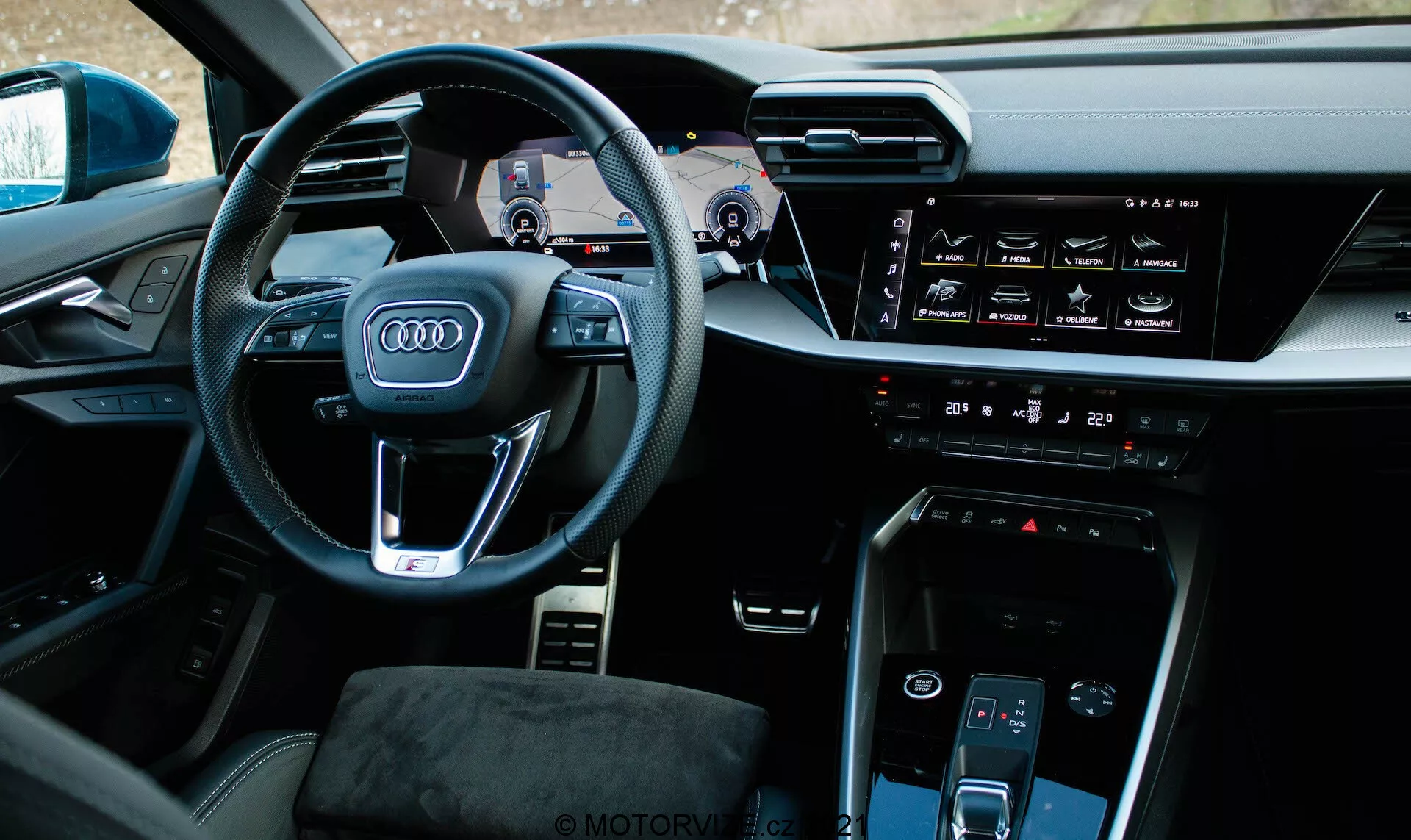 Vista interior de un Audi A3 Sportback 2020 desde un ángulo ligeramente descentrado a la izquierda centrada en el lado del conductor. El volante prominente presenta el icónico logotipo de los cuatro anillos de Audi, botones multifuncionales y paletas de cambio visibles. Detrás del volante, el panel de instrumentos digital, conocido como Audi Virtual Cockpit, muestra el velocímetro, tacómetro y mapa de navegación. La consola central incluye una pantalla táctil horizontal para infoentretenimiento, interfaz de control de clima, botón de arranque y parada del motor, otra pantalla táctil para el control del clima y un selector de marchas minimalista. El interior muestra materiales de alta calidad y un diseño tecnológicamente integrado con una sensación premium.
