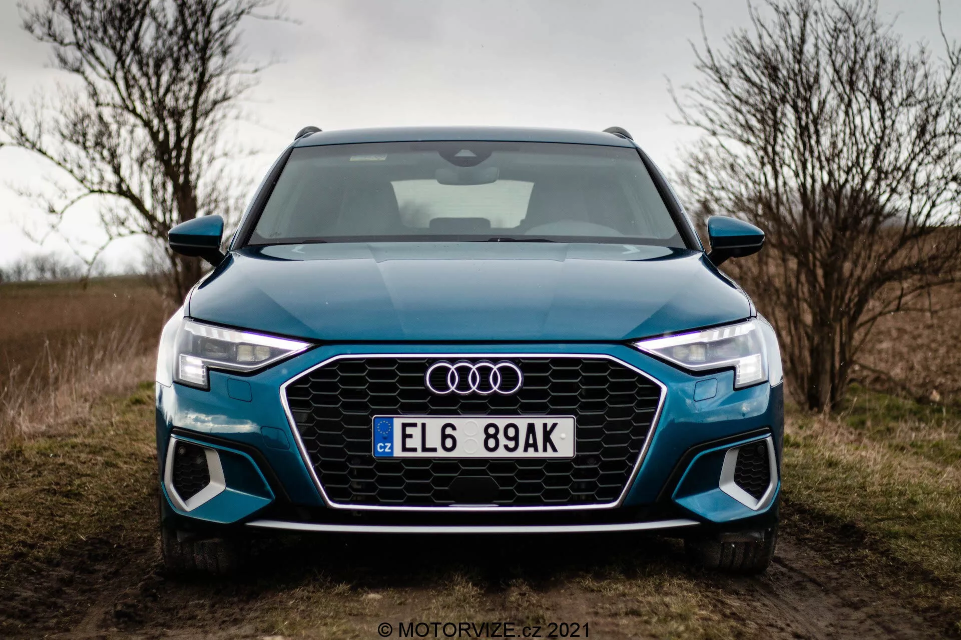 Vista frontal de un Audi A3 Sportback 2020 fotografiado desde un ángulo bajo. La imagen se centra en la parte frontal del automóvil, mostrando la parrilla octogonal Singleframe con inserciones en forma de panal, luces LED angulares y afiladas con distintivas luces diurnas, y entradas de aire grandes en el parachoques delantero esculpido. Los cuatro anillos de Audi están montados en el centro de la parrilla, con una matrícula de estilo europeo debajo. El coche, de un tono azul brillante, está aparcado al borde de un camino con césped y con un cielo nublado de fondo, lo que sugiere una estación otoñal o invernal.