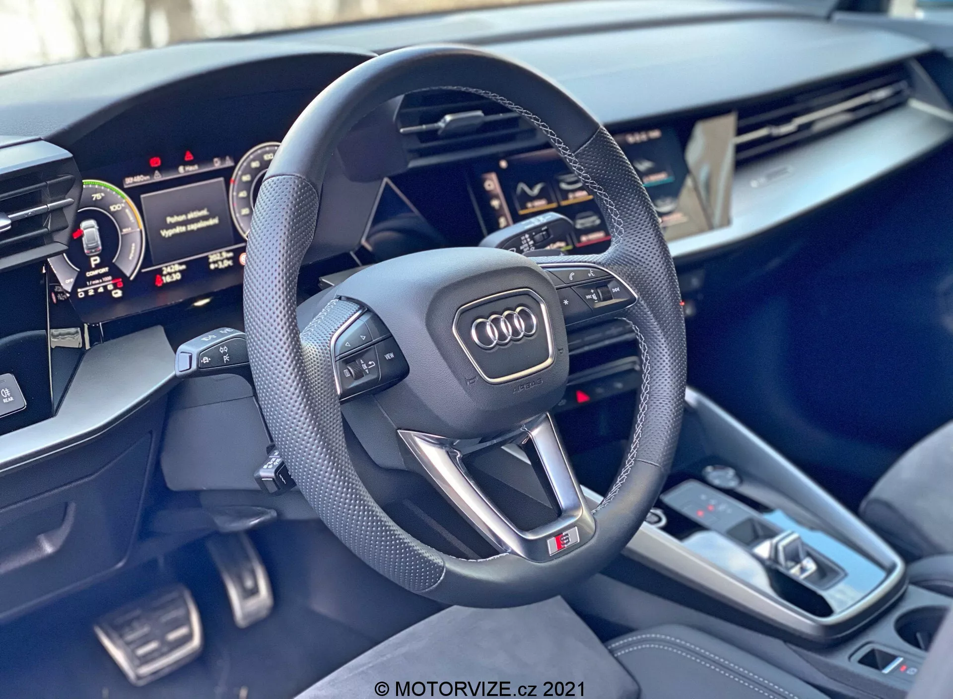 Vista interior de un Audi A3 Sportback 2020 centrada en el volante y la consola central. El volante deportivo de tres radios con parte inferior plana tiene empuñaduras de cuero perforado, botones multifuncionales, un emblema de Audi y una insignia 'S'. Detrás del volante se ve parcialmente el cuadro de instrumentos digital y a la derecha se muestra parcialmente el display táctil de la Interface Multimedia Central (MMI). La consola central incluye una palanca de cambios automática, múltiples botones, una almohadilla táctil, dos portavasos y pequeños compartimientos de almacenamiento. Los materiales del interior tienen detalles premium de metal cepillado y acabados de alta calidad, que enfatizan el lujo y la tecnología avanzada del coche.