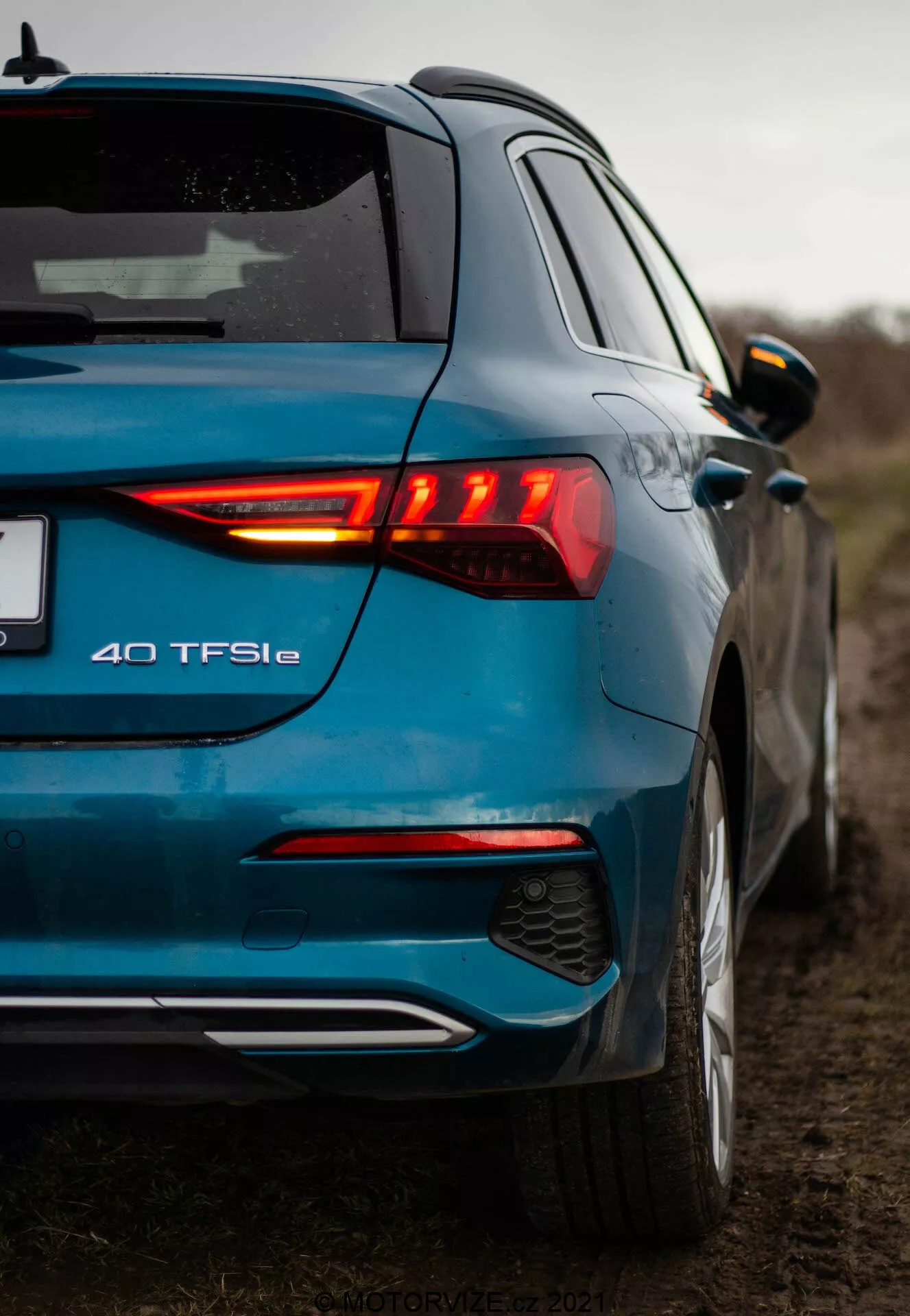 Una foto en ángulo bajo de un moderno Audi A3 2020 azul (A3 Sportback) tomada desde una vista de tres cuartos traseros, resaltando el grupo de luces traseras LED iluminadas, el emblema del modelo 40 TFSIe, el paragolpes trasero de doble tono con difusor integrado y sensores de estacionamiento, el acabado metálico de pintura, el alerón de techo, los espejos laterales del mismo color de la carrocería con indicadores integrados, y una insinuación de la puerta delantera y el perfil lateral. La fotografía enfatiza los elementos de diseño y los emblemas del coche en un entorno rural con cielo nublado y poca profundidad de campo.
