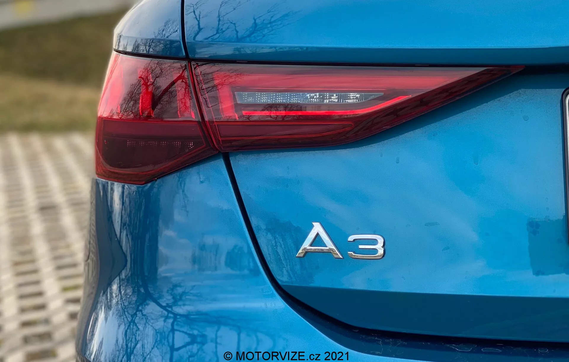 Esta imagen presenta una toma en primer plano y bajo ángulo de la esquina trasera izquierda de un Audi A3 Sportback 2020 azul. La fotografía enfatiza el diseño elegante de la luz trasera, que tiene una orientación horizontal afilada que se extiende hacia el lateral del vehículo, y la designación del modelo 'A3' en cromo justo debajo. Los elementos LED dentro de la luz trasera resaltan la tecnología avanzada de iluminación del coche. La pintura reluciente y la limpieza general del modelo muestran un detallado de automóvil de alta calidad, con un fondo ligeramente desenfocado que sugiere un entorno al aire libre. La marca de agua '© MOTORVIZE.cz 2021' indica la propiedad y el origen de la imagen.