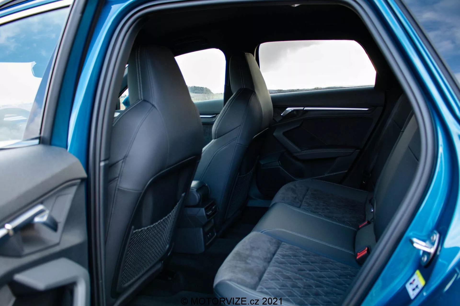 Toma interior de un Audi A3 Sportback 2020 visto desde el lado trasero del pasajero, mostrando asientos espaciosos para tres personas con reposacabezas y cinturones de seguridad, tapizados en tela oscura y con un patrón. Los asientos delanteros con diseño ergonómico son parcialmente visibles. Los elementos visibles del panel de la puerta incluyen un tirador y posibles controles. La imagen bien iluminada resalta la sensación elegante y premium con materiales y detalles de diseño de calidad.