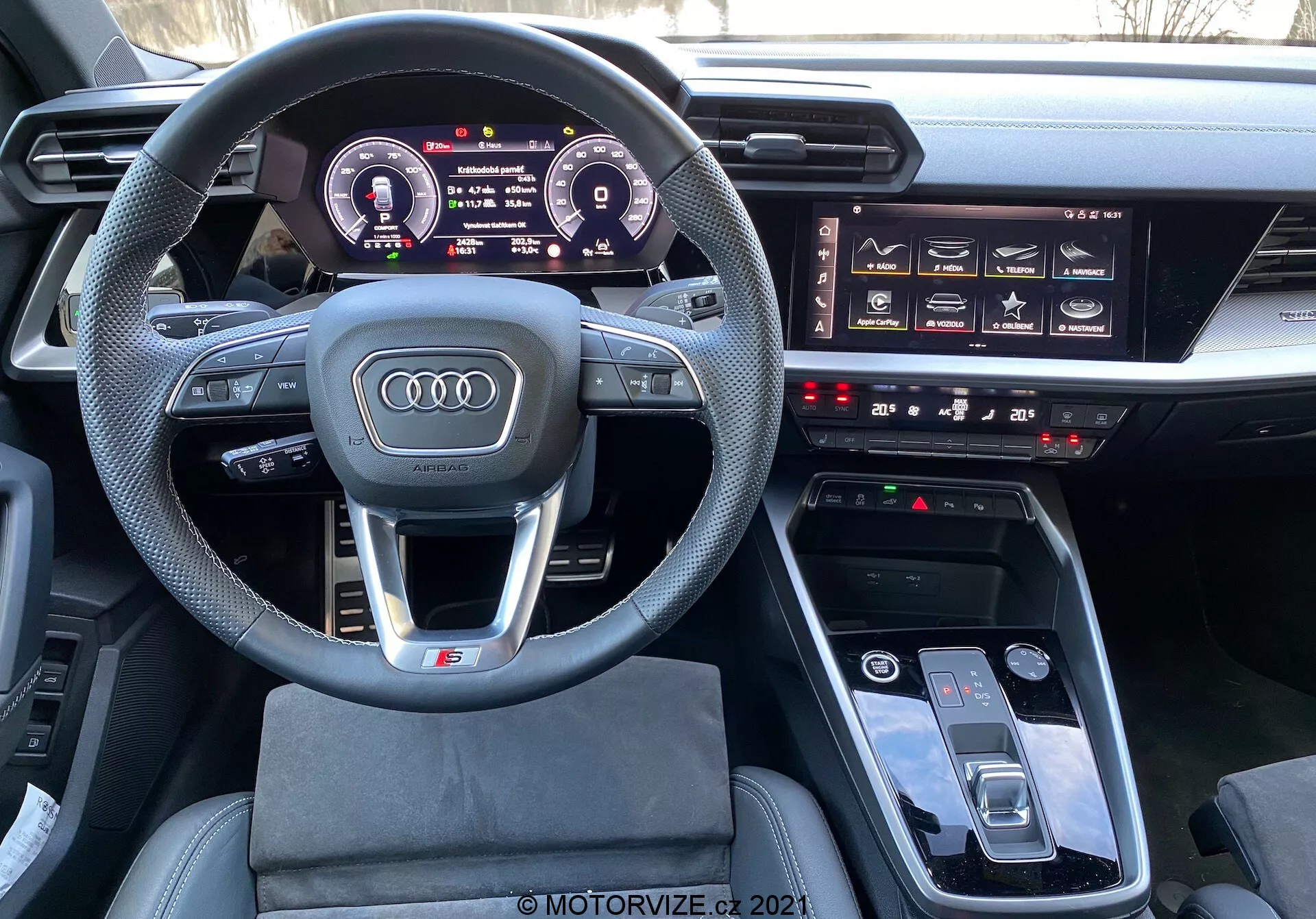 Vista interior del Audi A3 Sportback 2020 desde el asiento del conductor que muestra el volante deportivo multifuncional de cuero con diseño de fondo plano, panel de instrumentos digital Audi Virtual Cockpit, sistema de infoentretenimiento prominente, botones táctiles para el control climático y panel táctil MMI en la consola central, indicando un interior de automóvil moderno, lujoso y tecnológicamente avanzado.