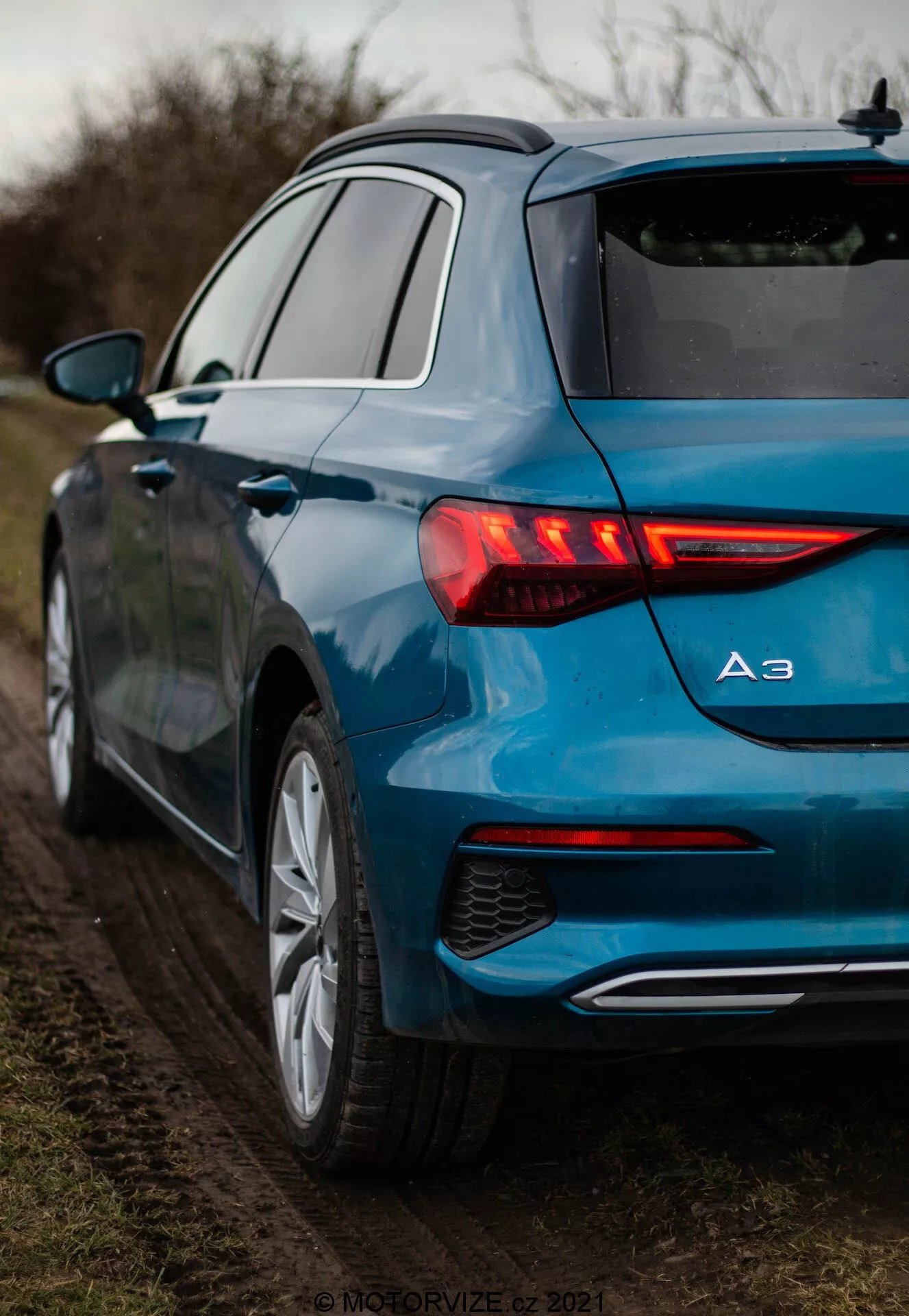 Vista detallada de un Audi A3 2020 Sportback azul metálico desde una perspectiva de tres cuartos trasera, destacando sus luces traseras LED iluminadas con señales de giro dinámicas, la insignia del modelo que muestra 'A3', ruedas de aleación multiradio y una salida de escape única. El coche está aparcado en un borde de carretera sin pavimentar rodeado de vegetación, bajo una iluminación suave y difusa.