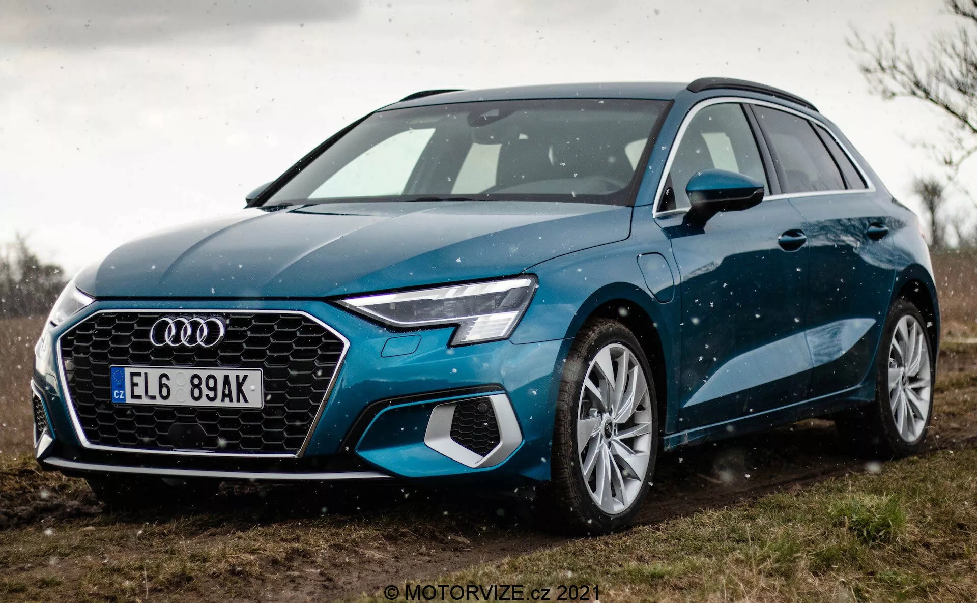 Vista en ángulo bajo de un Audi A3 Sportback 2020 azul, destacando la rejilla frontal y el diseño de los faros LED. La perspectiva cercana se centra en el lado izquierdo frontal del coche, mostrando su rejilla hexagonal con el prominente emblema de Audi, sensores integrados para características ADAS, grandes llantas de aleación multiradio y espejos laterales elegantes. El coche está aparcado sobre una superficie no pavimentada con vegetación al fondo, sugiriendo un entorno rural. Gotitas de agua sutiles en la carrocería del coche acentúan sus líneas y color.