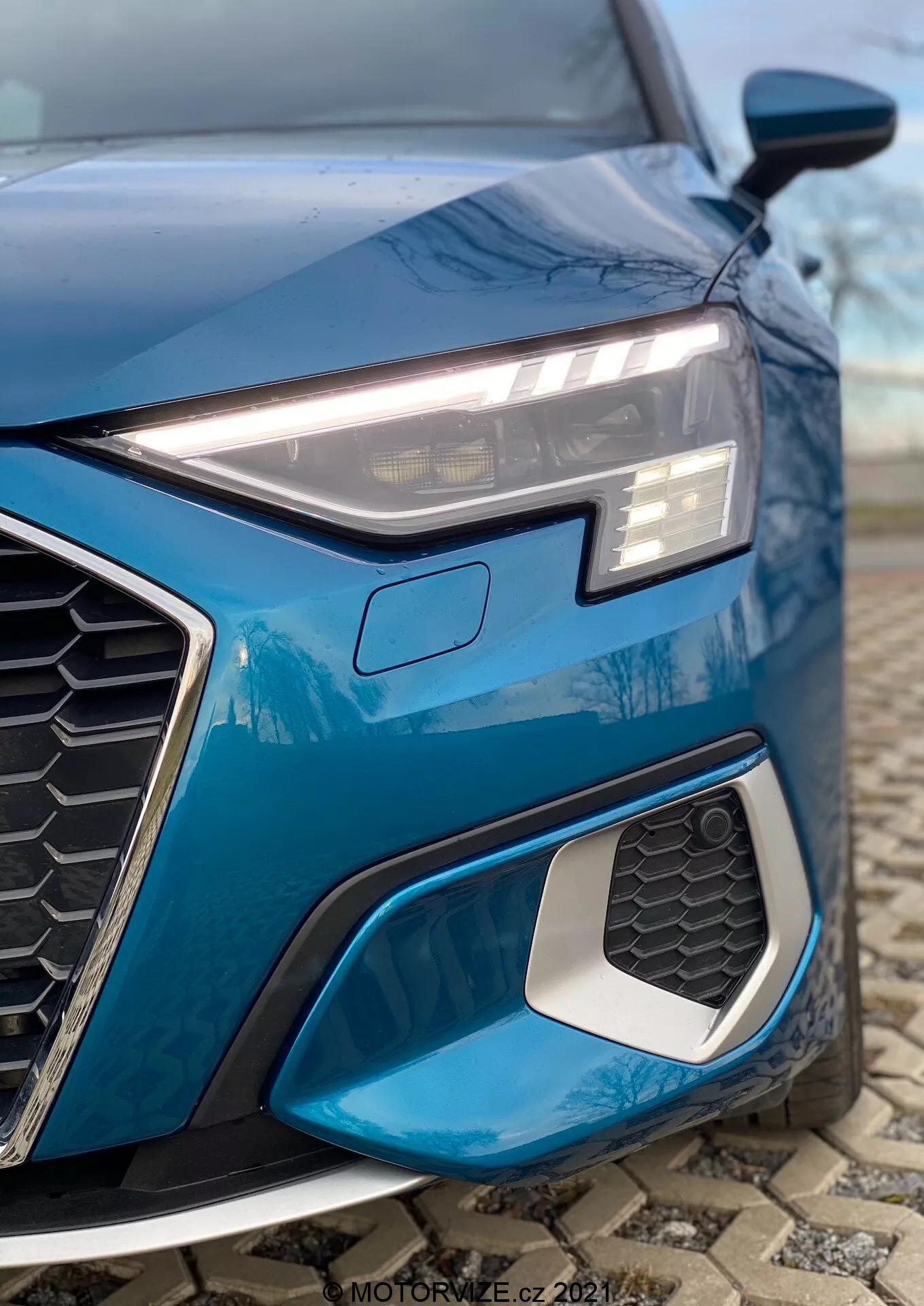 Primer plano del cuarto delantero izquierdo de un Audi A3 2020 azul (A3 Sportback), destacando el elegante faro LED angular con luces diurnas, la prominente parrilla hexagonal con diseño de panal y la entrada de aire con borde metálico. La toma a bajo ángulo resalta los elementos del diseño deportivo, el diseño geométrico intrincado de las luces antiniebla y el fondo desenfocado para una mayor atención al detalle.