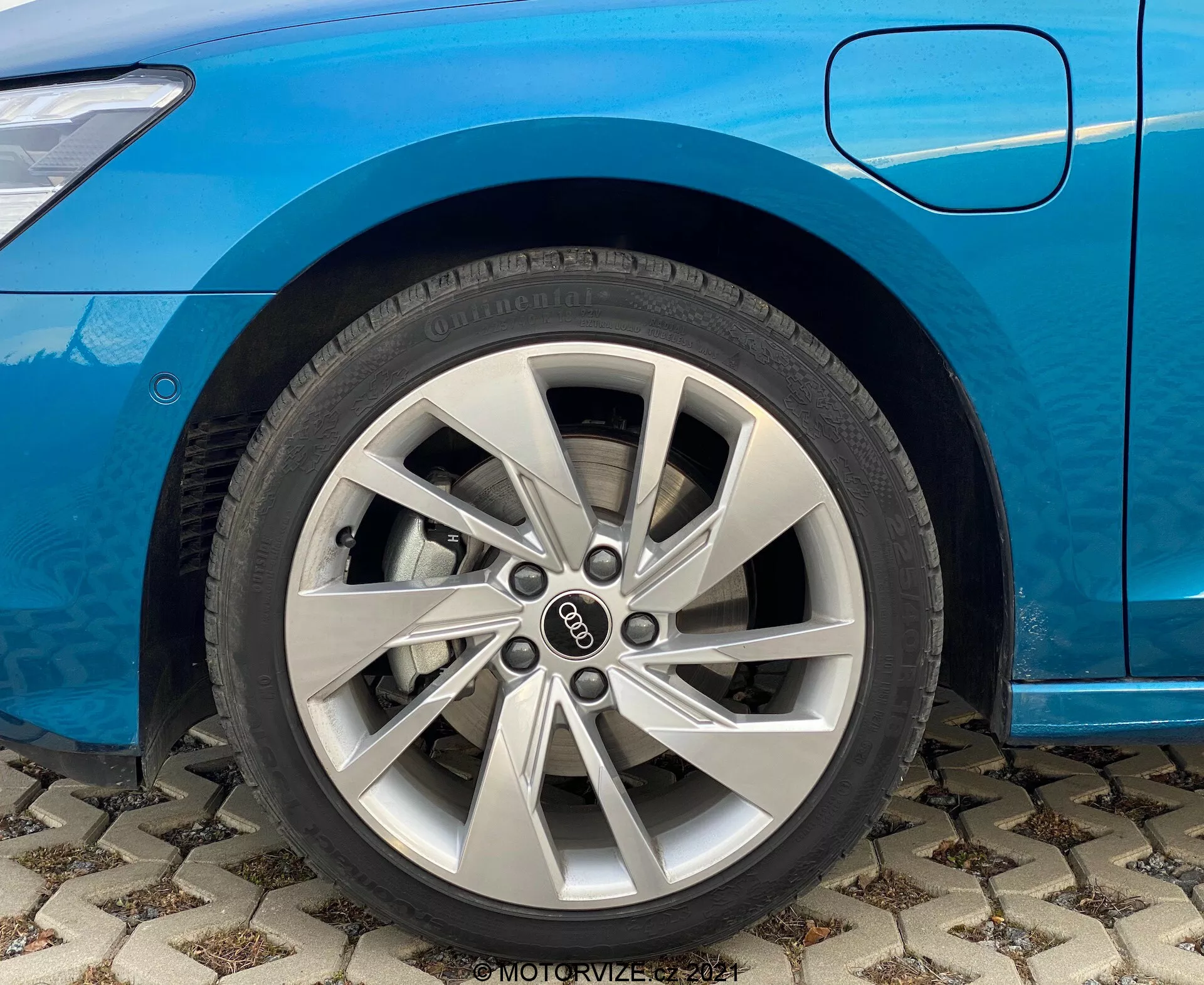 Primer plano de la sección de la rueda delantera de un Audi A3 2020 azul (A3 Sportback). Vista enfocada en la llanta de aleación plateada de varios radios en el lado del pasajero, mostrando también el guardabarros delantero y el borde inferior de la puerta del pasajero. El coche está aparcado sobre una superficie pavimentada con un diseño, con detalles visibles de la banda de rodadura y de la marca del neumático. Notable para discusiones sobre diseño automotriz y técnicas de fabricación de ruedas. Incluye una marca de agua que dice 'MOTORVIZE 2021.'