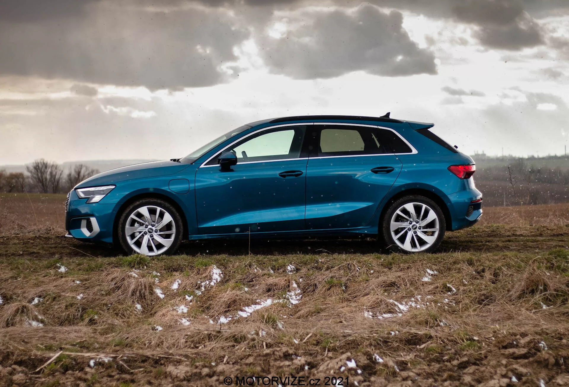 Vista de perfil lateral de un Audi A3 Sportback azul 2020 estacionado en un campo desigual con parches de hierba y posiblemente restos de nieve o hielo, con un cielo nublado, destacando el diseño dinámico del coche con una línea de techo suavemente inclinada, llantas de aleación multirradio, vidrios ligeramente tintados, espejos laterales del color de la carrocería con señales de giro integradas, y faros y luces traseras LED afilados.
