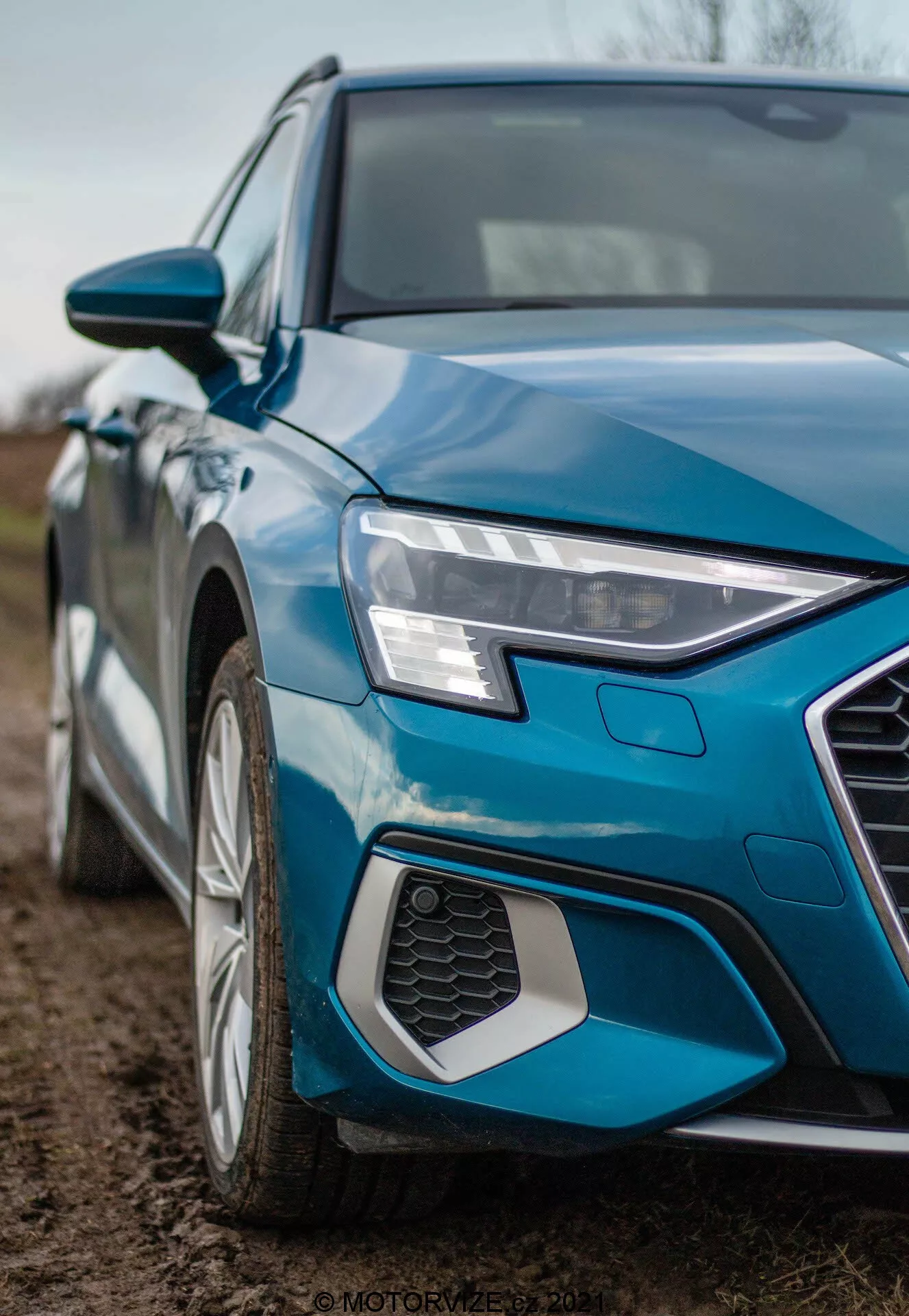 Imagen detallada en ángulo oblicuo del Audi A3 Sportback 2020, enfocándose en el cuadrante delantero izquierdo. La fotografía en ángulo bajo captura el faro delantero izquierdo afilado y angular con luces diurnas LED, parte de la parrilla hexagonal de panal, el guardabarros delantero izquierdo y un segmento de la puerta del conductor. También es visible la llanta de aleación con diseño de múltiples radios, la pintura azul metálica y el espejo lateral del color de la carrocería con señales de giro integradas, acentuando el diseño moderno y las características tecnológicas.