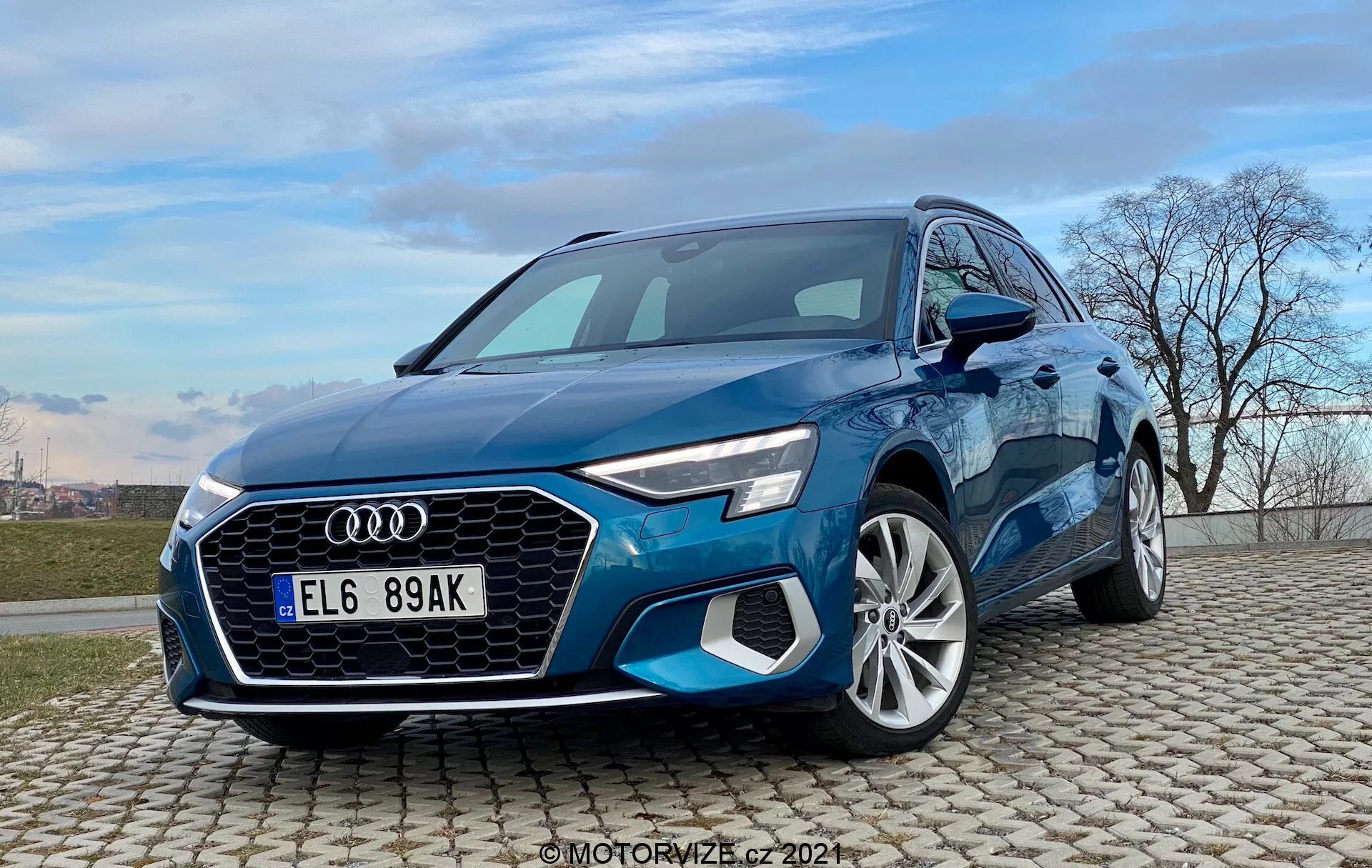 Vista de tres cuartos delantera de un Audi A3 Sportback azul 2020 capturada desde un ángulo bajo. La imagen muestra el aspecto dinámico e imponente del coche con la distintiva parrilla Singleframe, forma hexagonal con lamas verticales, faros LED afilados y entradas de aire en el parachoques delantero. El capó esculpido lleva hacia el parabrisas, revelando contornos aerodinámicos y líneas de carrocería. El perfil lateral presenta guardabarros definidos sobre ruedas de aleación multirradio y pilares A inclinados con espejos laterales elegantes. El coche está posicionado sobre un pavimento adoquinado bajo un cielo azul claro con nubes y una línea de árboles desnudos, sugiriendo un contexto promocional o de revisión de coches.