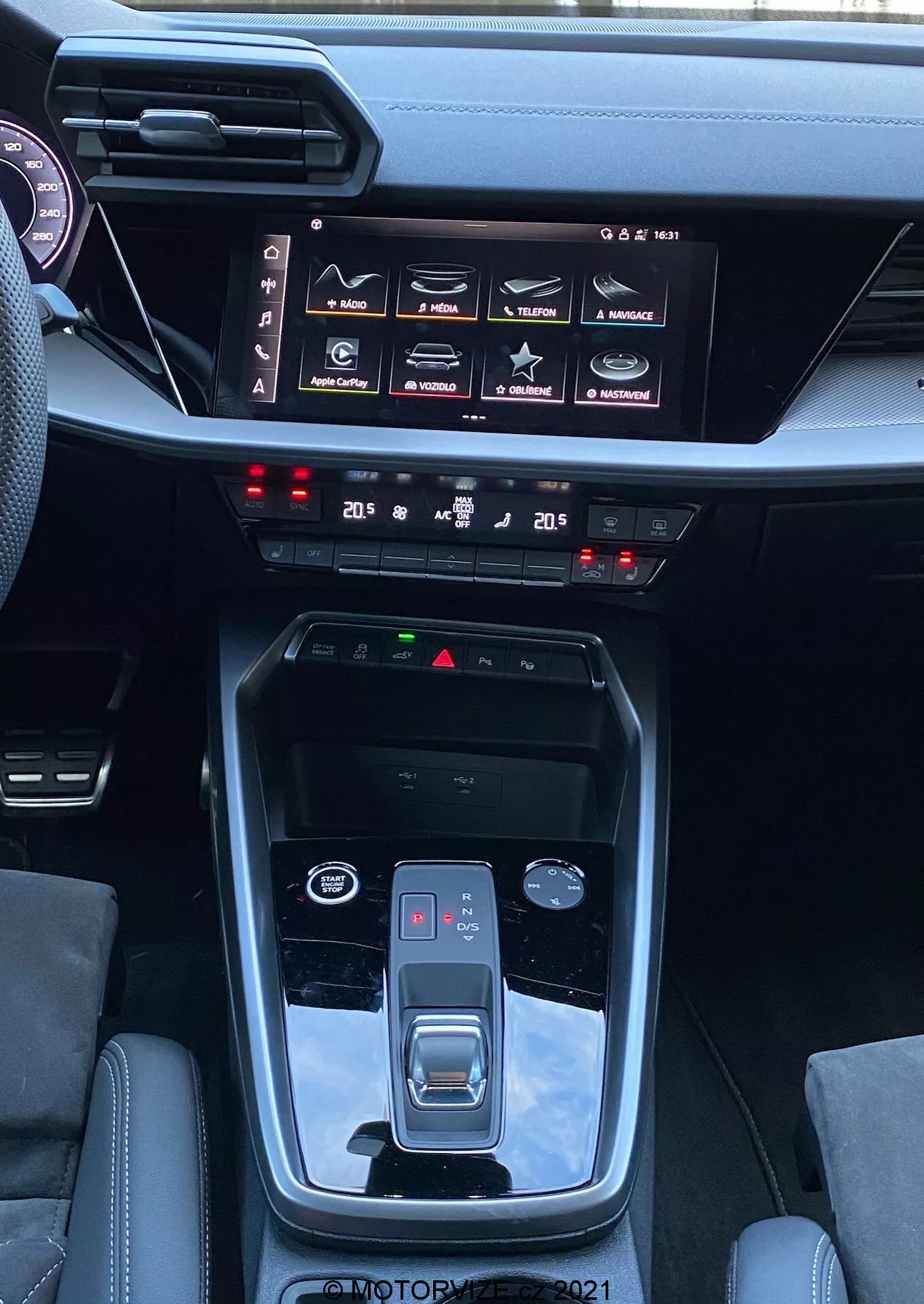 Vista interior del Audi A3 2020 Sportback que muestra la consola central y el tablero, destacando características como la palanca de cambios automática con botón de estacionamiento, freno de mano electrónico, selector de modos de conducción, acentos metálicos, sistema de infoentretenimiento con pantalla táctil y varias configuraciones del coche, control de clima dual, botones de características de seguridad y diseño elegante y moderno del tablero.