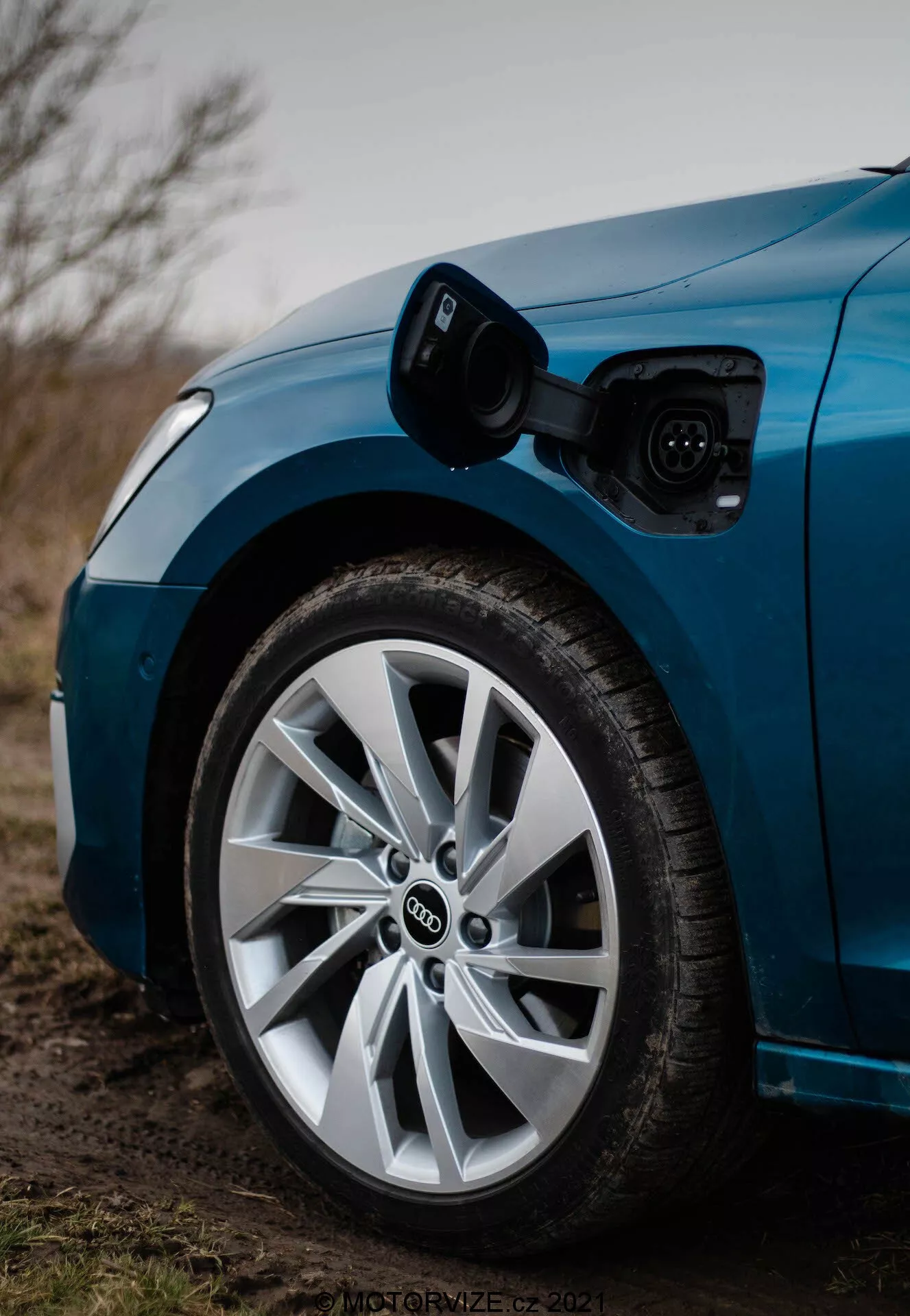 Primer plano del cuarto delantero del lado del conductor de un Audi A3 2020 Sportback azul, destacando la rueda y el puerto de carga. La imagen, tomada desde un ángulo bajo hacia el frente, muestra el puerto de carga abierto sobre la rueda delantera, con un cable de carga conectado. La poca profundidad de campo enfoca nítidamente en la rueda y el puerto de carga, revelando el logotipo de anillos superpuestos característico de Audi y el diseño de rueda multirradio con un neumático de perfil bajo, todo bajo una iluminación ligeramente tenue, posiblemente de anochecer.