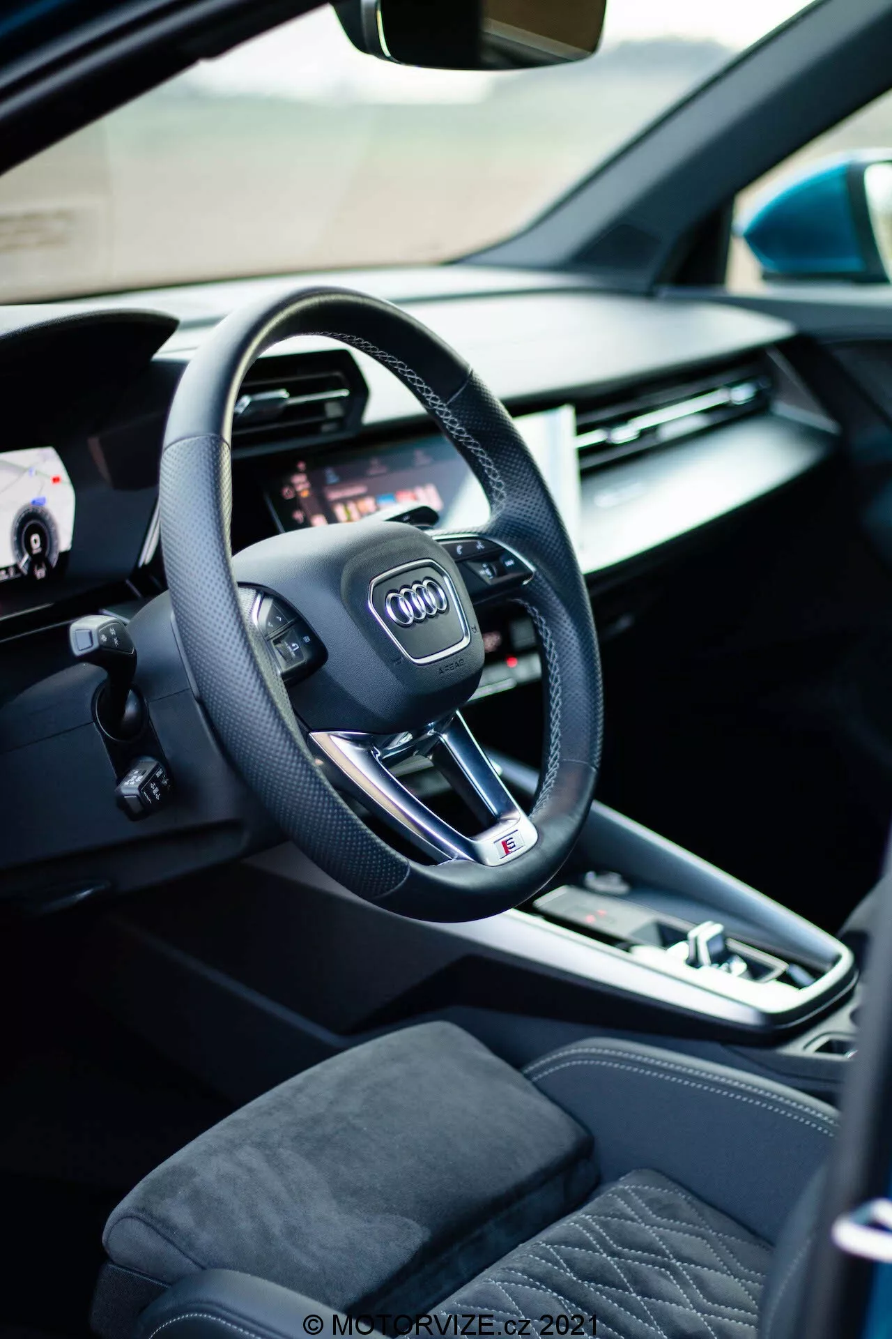 Vista interior de un Audi A3 Sportback 2020, centrada en el lado del conductor con un volante moderno de tres radios que muestra el logotipo de Audi, botones multifunción integrados y un diseño de base plana que indica un rendimiento deportivo. Detrás del volante, se puede ver parte de un panel de instrumentos digital o cabina virtual, que ofrece opciones de visualización personalizables para diversas funciones del coche. A la izquierda, una puerta de coche abierta revela controles de ventana eléctrica y ajustes de memoria de los asientos, enfatizando la conveniencia y la personalización. La consola central incluye un selector de transmisión automática, un botón de freno de estacionamiento electrónico y un panel táctil/controlador para el sistema de infoentretenimiento, lo que destaca la participación del conductor. Los asientos visibles están tapizados con materiales premium con un diseño acolchado, lo que añade a la sensación de lujo del interior.