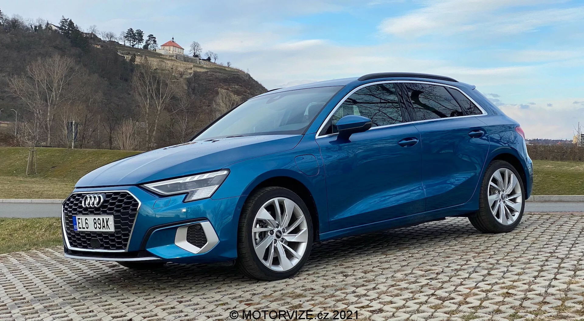 Vista a tres cuartos desde el frente de un Audi A3 Sportback 2020 azul brillante en adoquines, mostrando elementos de diseño distintivos como faros LED, parrilla prominente, llantas de aleación de múltiples radios y sistemas de seguridad avanzados. Capturado con un fondo de colinas con hierba y cielo parcialmente nublado.