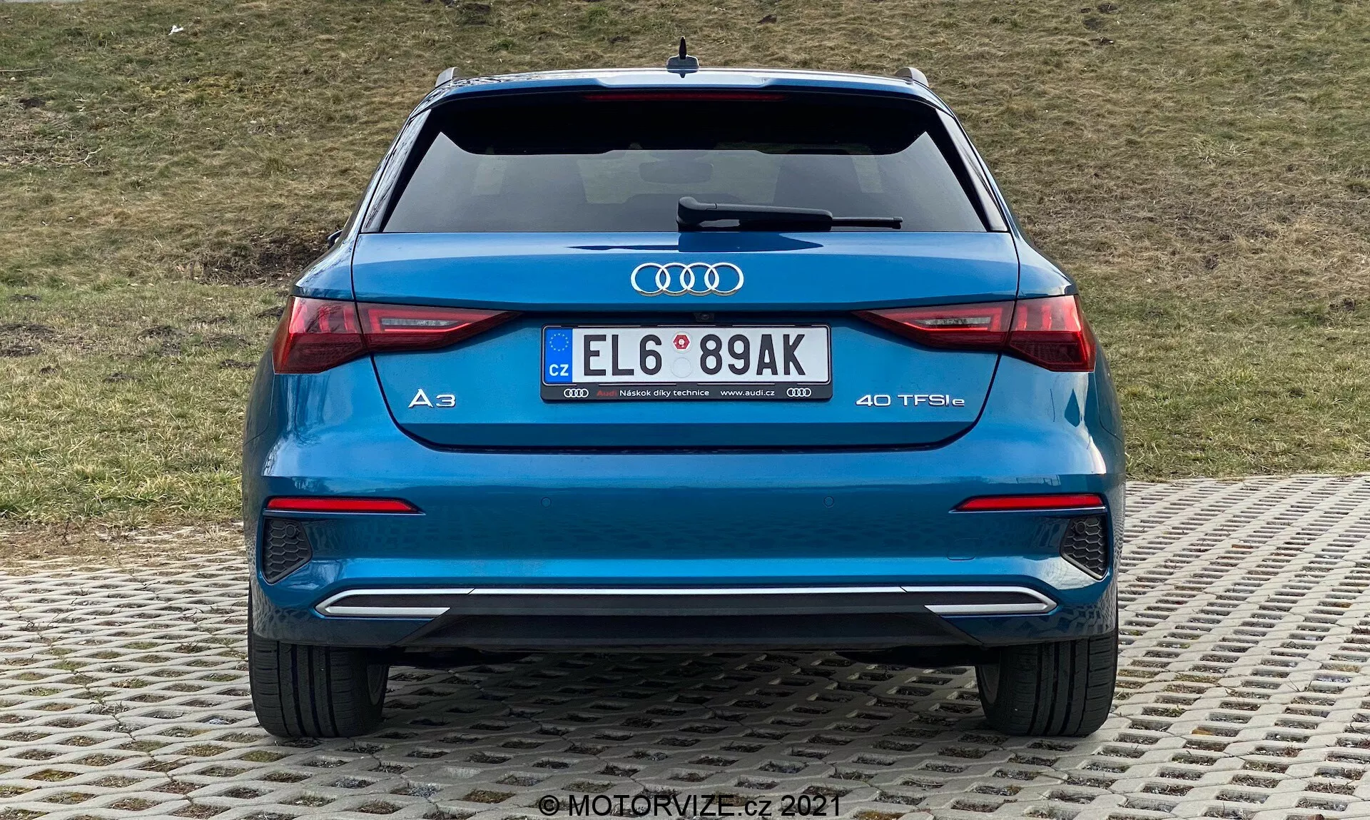 Vista trasera de un Audi A3 2020 Sportback azul, mostrando el parachoques trasero, maletero, luces traseras, tubos de escape y el parabrisas trasero. Logotipo de Audi y designación del modelo 'A3' con la insignia '40 TFSI' que indica un motor de gasolina turboalimentado. Luces traseras LED con diseño horizontal elegante, dobles tubos de escape, parachoques trasero con elemento difusor y sensores de aparcamiento, y matrícula de estilo europeo. Fondo con una pendiente cubierta de hierba.