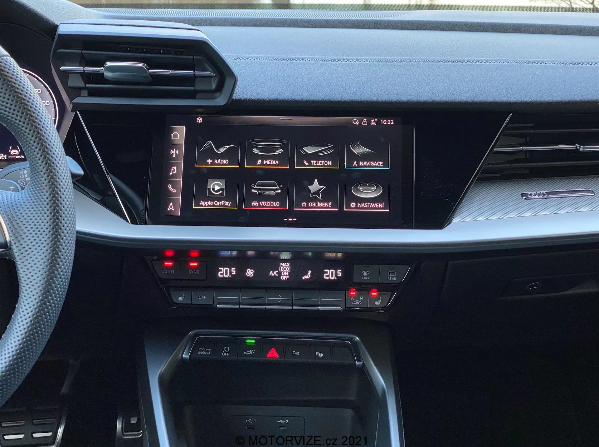Vista del tablero interior del Audi A3 Sportback 2020, destacando su avanzado sistema de infoentretenimiento y controles climáticos. La imagen capta una gran pantalla táctil horizontal con características como navegación, medios, conectividad telefónica y configuraciones del coche, indicando capacidades integrales de infoentretenimiento incluyendo Apple CarPlay y Android Auto. Debajo de la pantalla, los controles de clima digital de doble zona muestran configuraciones a 20 grados Celsius para conductor y pasajero. El tablero también incluye un área de carga inalámbrica y delgadas rejillas de ventilación horizontales, ejemplificando el diseño moderno de interiores automotrices. La foto tiene una marca de agua 'MOTORVIZE.CZ 2021'.