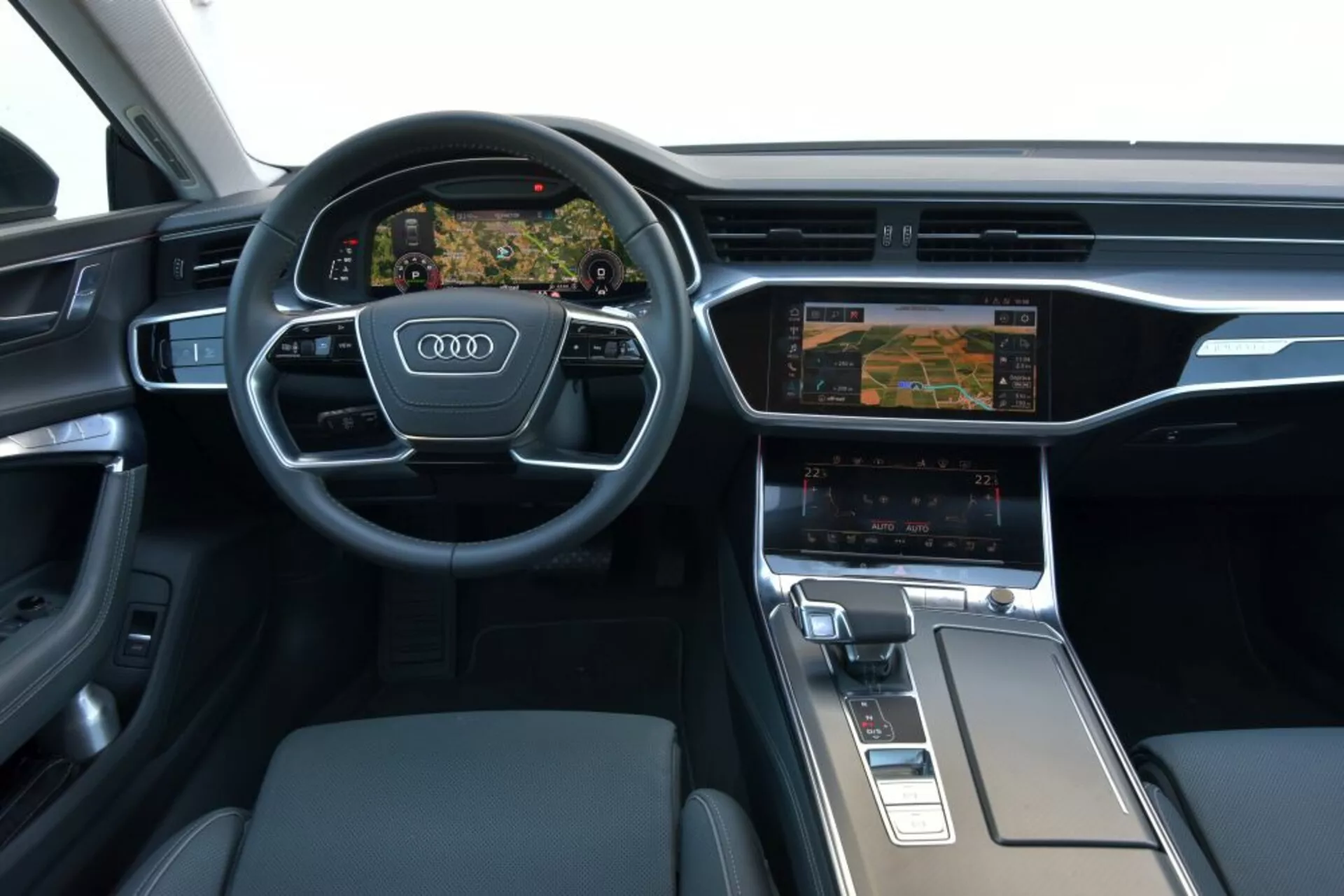 Vista interior del Audi A7 Sportback 2018 enfocada en el tablero de mandos y la zona de la consola central del conductor, mostrando un volante multifunción de tres radios con el logo de Audi, un cuadro de instrumentos digital Virtual Cockpit que muestra la navegación, sistema de infoentretenimiento con doble pantalla con configuraciones de control climático, selector de marchas ergonómico y materiales premium como metal cepillado y cuero.