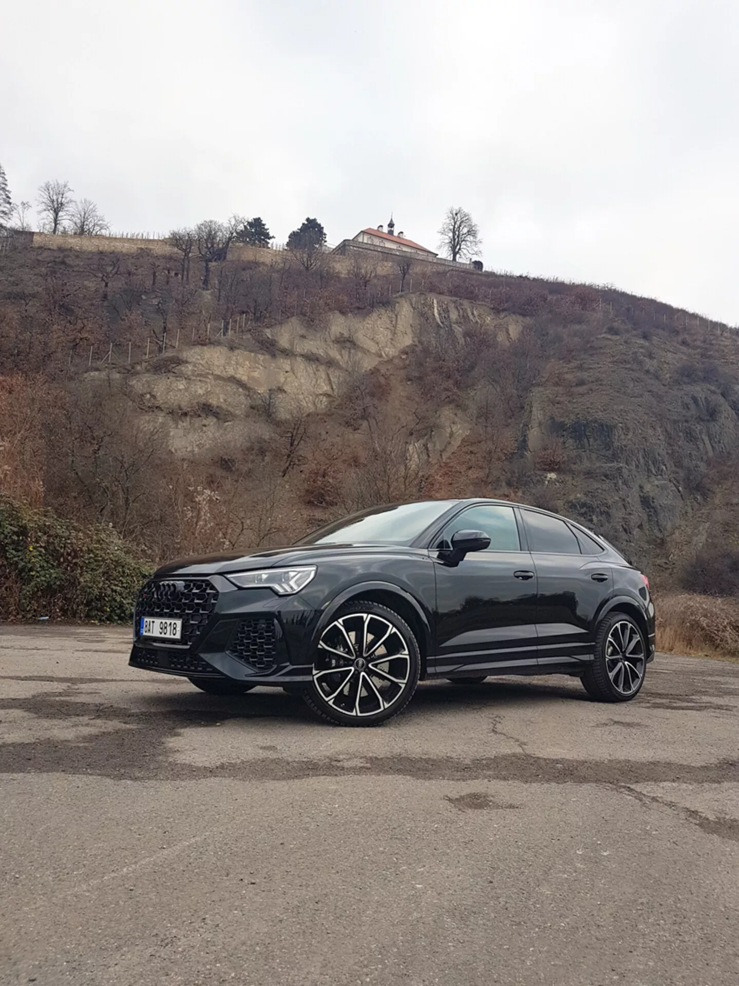 Audi Q3 RS 2015 negro, un vehículo utilitario deportivo estacionado en un área pavimentada con fondo de colina rocosa, mostrando sus grandes ruedas de aleación de múltiples radios, faros elegantes equipados con LED y líneas de carrocería deportivas bajo un cielo nublado.