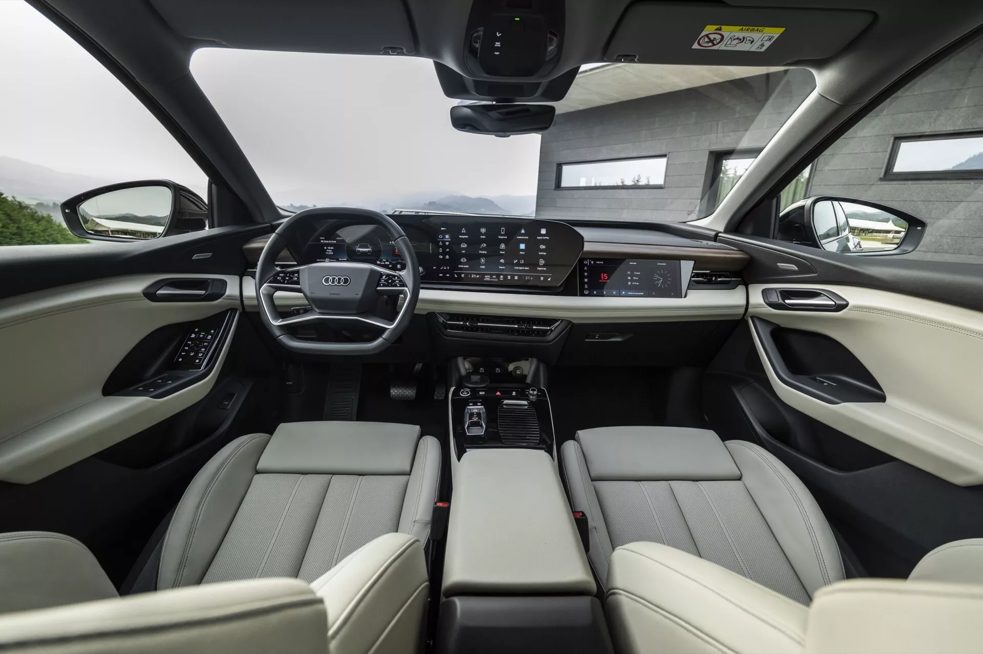 Vista interior del Audi Q6 e-tron 2024 desde la perspectiva del asiento trasero, enfocándose en la fila delantera. Los asientos delanteros de cuero claro presentan reposacabezas ajustables y controles electrónicos. El tablero de instrumentos muestra un panel digital expansivo y una gran pantalla táctil central para el sistema de infoentretenimiento, rodeada de botones de control para el clima y los modos de conducción. La consola central incluye portavasos, espacio de almacenamiento y un selector de cambio automático. El volante está equipado con múltiples controles y el techo panorámico sugiere abundante luz natural. En conjunto, la imagen destaca tecnologías avanzadas y características automotrices de lujo.