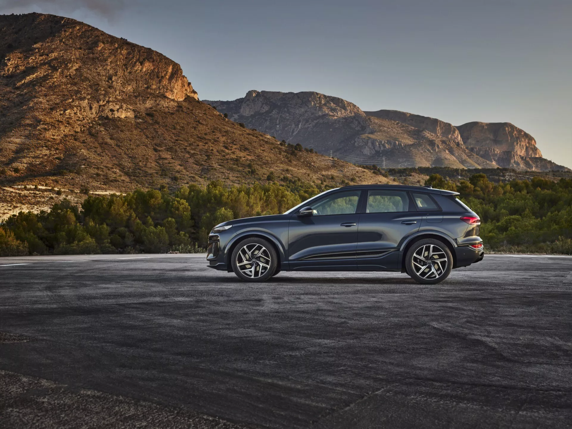 Un SUV moderno, que se cree que es el Audi Q6 e-tron 2024, estacionado al aire libre con un fondo montañoso, capturado en una vista trasera de tres cuartos desde un ángulo elevado. El color de la carrocería metálico oscuro contrasta con el entorno natural, destacando líneas elegantes y contornos aerodinámicos. Los detalles visibles incluyen luces traseras LED, grandes llantas de aleación con diseño de múltiples radios, un sistema de escape doble, ventanas tintadas, un techo solar panorámico y molduras protectoras a lo largo de los bordes inferiores. La ventana trasera está ligeramente inclinada con un alerón trasero y espejos laterales de tamaño considerable posiblemente con intermitentes integrados, realzando el atractivo deportivo y lujoso del vehículo.